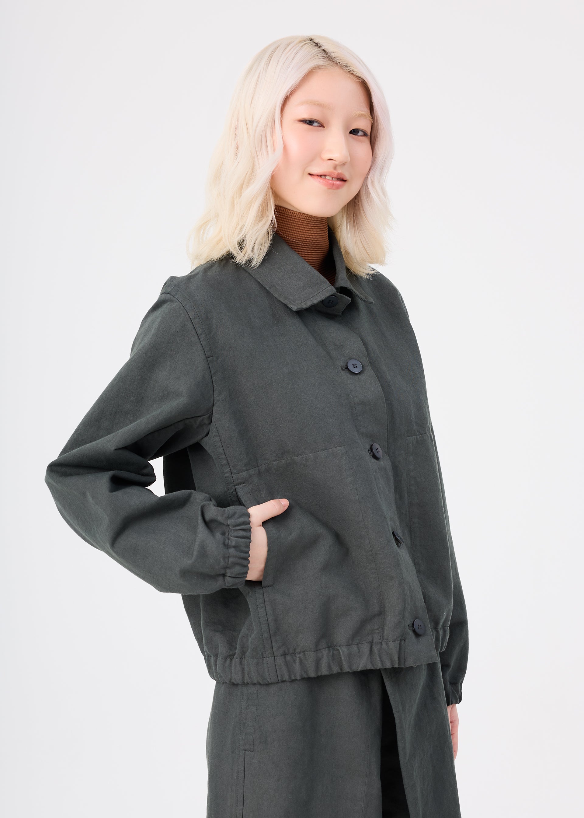 Zoomed view A person with light blonde hair is wearing the me ISSEY MIYAKE PINA CLOTH garment-dyed jacket and matching pants, standing against a plain white background and smiling gently with one hand in a pocket.