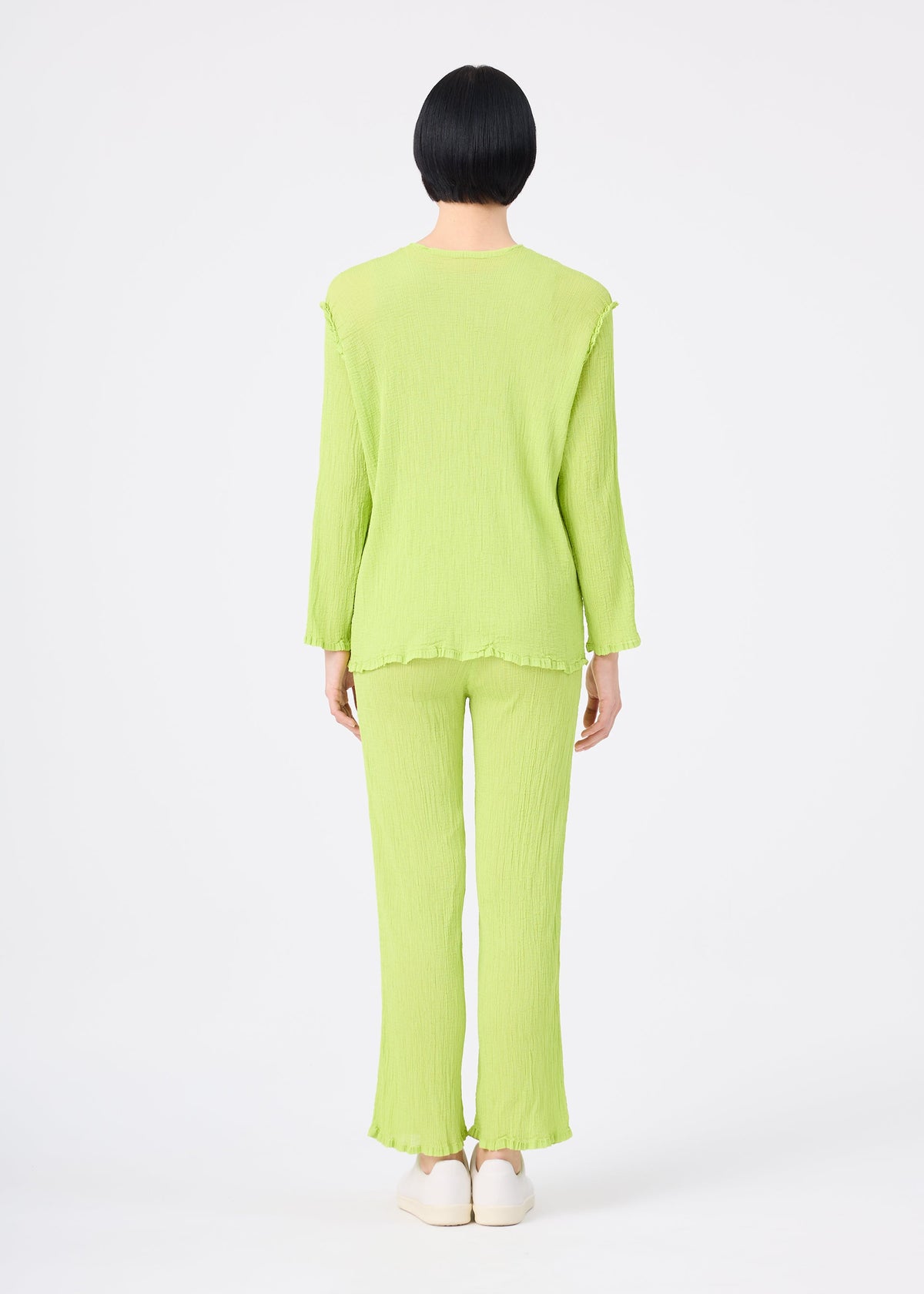 A person with short black hair faces away, wearing the me ISSEY MIYAKE SEE-THROUGH CREPE COLOR set—a light green, long-sleeve, loose-fit top and matching crinkled pants—paired with white shoes against a plain white background. view 6 of 6