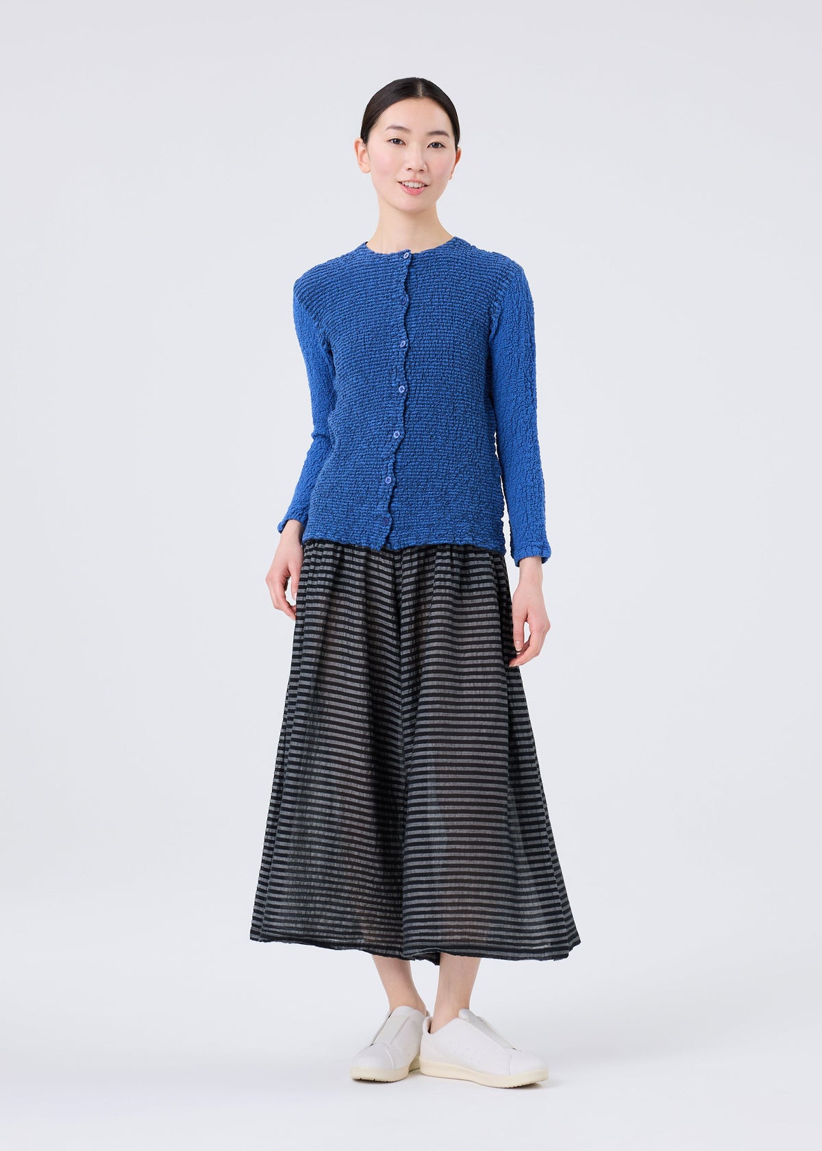 A woman stands facing forward against a plain light background, wearing a blue textured cardigan, white slip-on shoes, and a black and gray midi skirt made from soft STRIPE CAULIFLOWER fabric by me ISSEY MIYAKE for cotton comfort. view 6 of 8