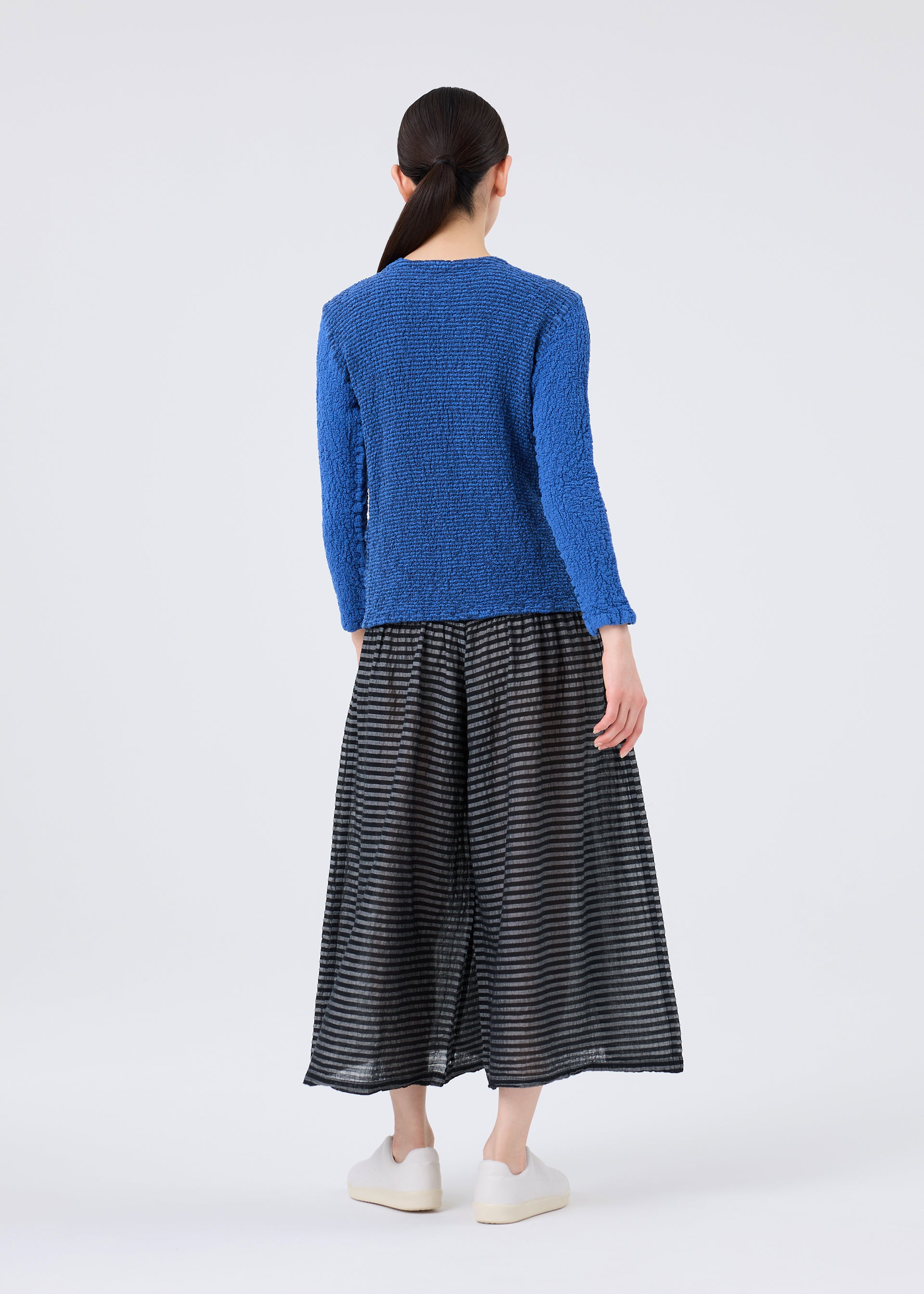 Zoomed view A woman stands with her back to the camera against a plain background, wearing a textured, vibrant blue long-sleeved top crafted for cotton comfort. She pairs it with the me ISSEY MIYAKE STRIPE CAULIFLOWER midi skirt—a loose-fitting, semi-sheer piece featuring black-and-grey stripes—and simple white slip-on sneakers.