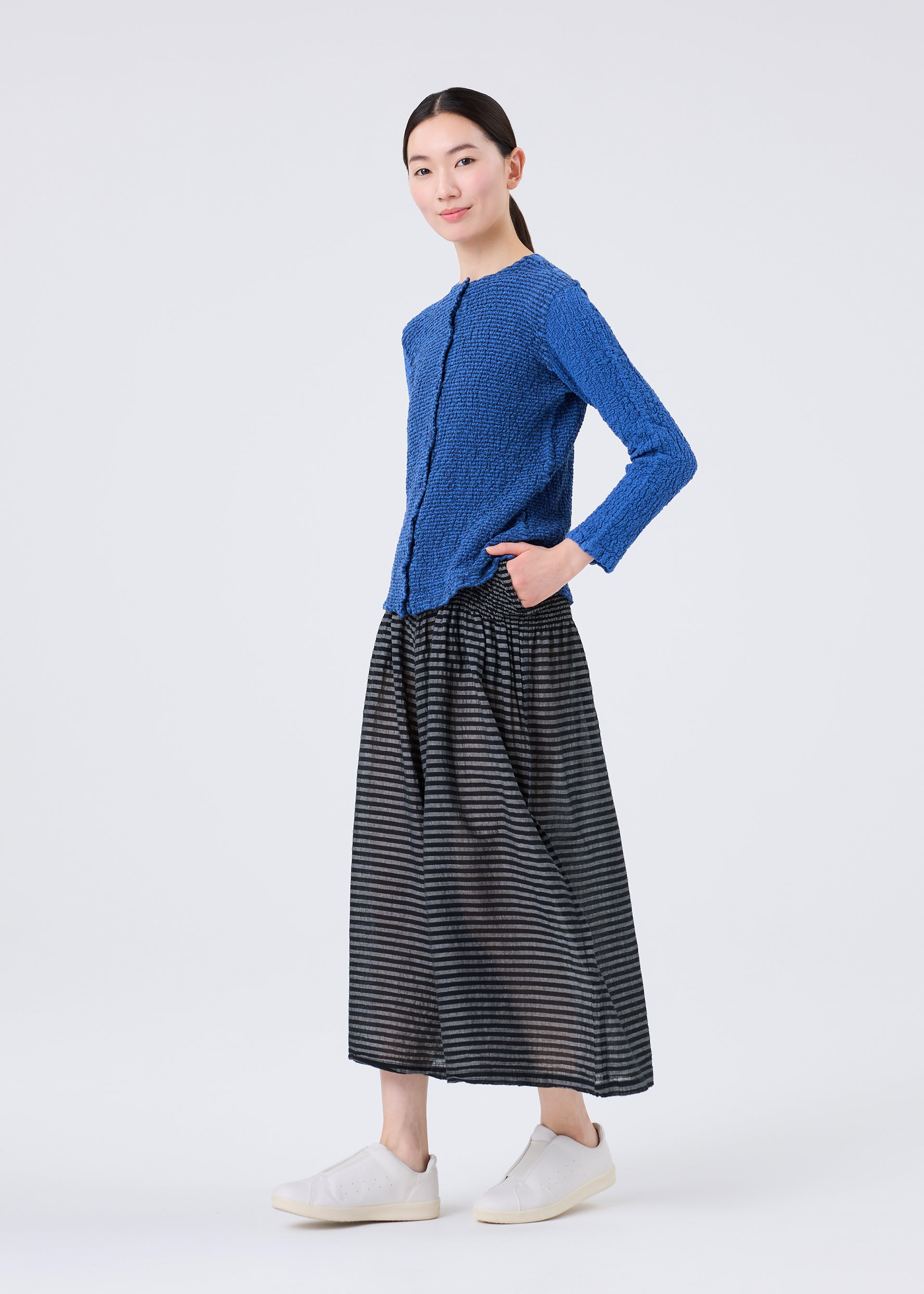 Zoomed view A woman stands against a white background, wearing a textured blue long-sleeve top, white slip-on shoes, and a black and gray skirt from me ISSEY MIYAKE’s STRIPE CAULIFLOWER collection. She exudes cotton comfort with one hand in her pocket and a slight smile.