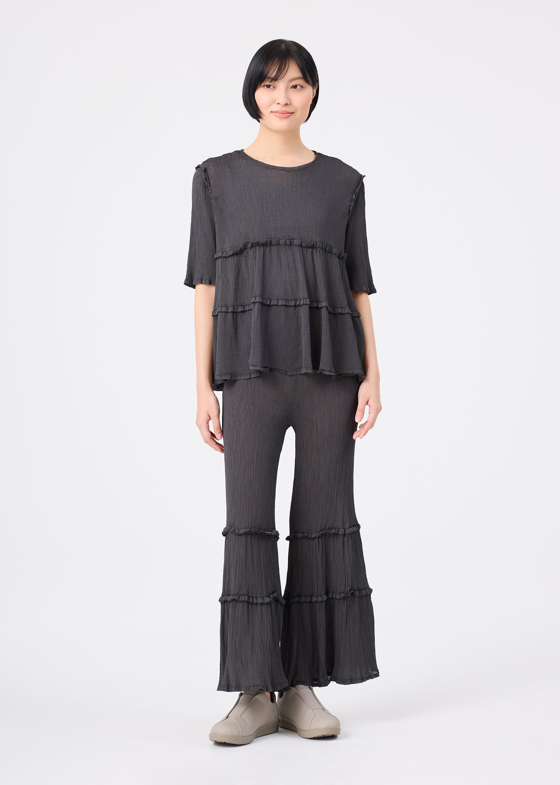Zoomed view A person stands against a plain white background wearing the FLARE SEE-THROUGH CREPE set from me ISSEY MIYAKE, featuring a dark, layered ruffle top and matching flared pants with ruffle details in garment-dyed fabric, paired with light-colored shoes.