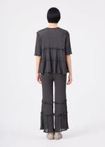 A person with short black hair stands facing away, wearing the me ISSEY MIYAKE FLARE SEE-THROUGH CREPE ensemble—a loose-fitting, dark gray, short-sleeved top and matching tiered pants featuring ruffle details—crafted from upcycled natural dyes and garment-dyed crepe fabric. The outfit is paired with beige shoes against a plain white background.