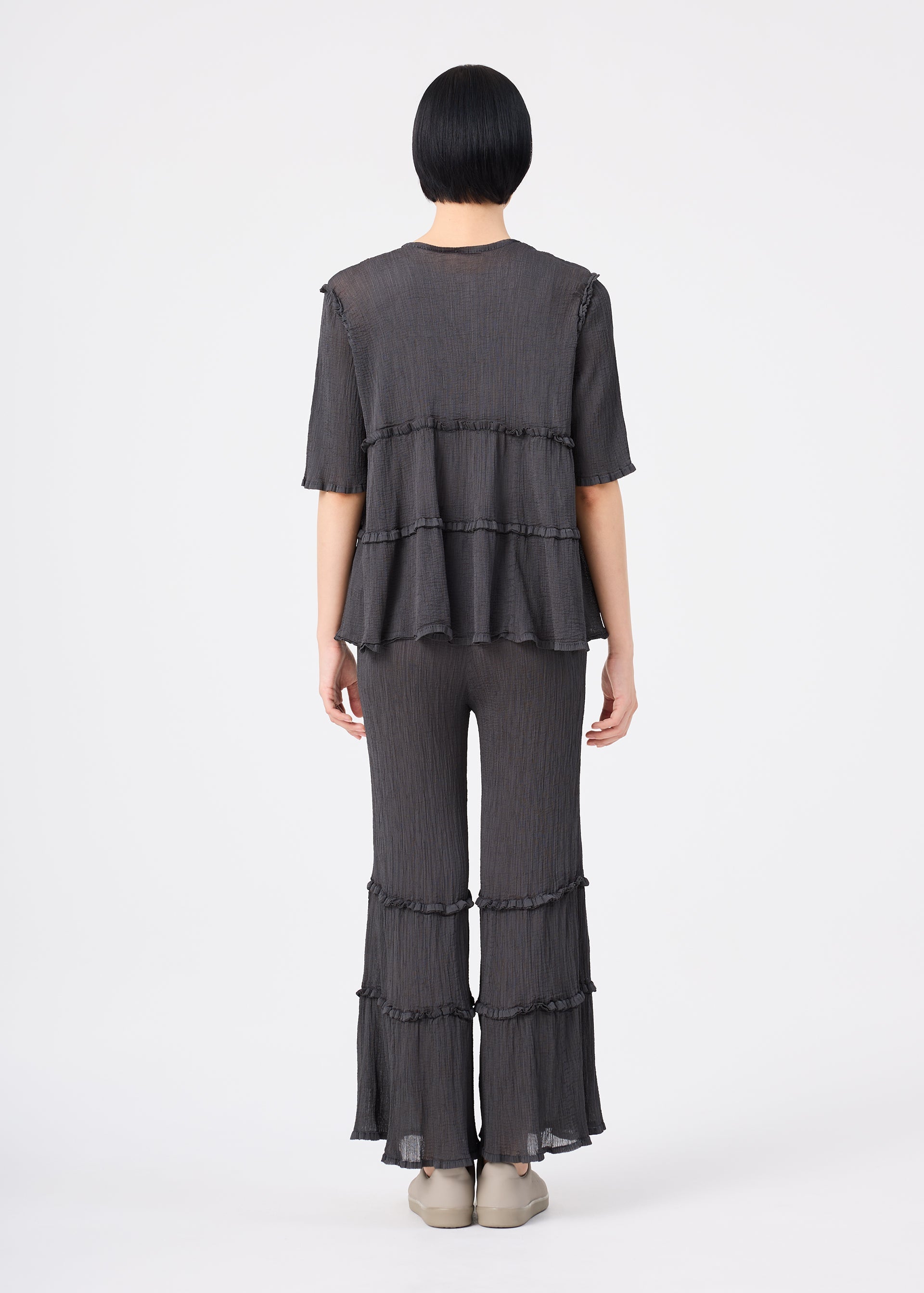 Zoomed view A person with short black hair stands facing away, wearing the me ISSEY MIYAKE FLARE SEE-THROUGH CREPE ensemble—a loose-fitting, dark gray, short-sleeved top and matching tiered pants featuring ruffle details—crafted from upcycled natural dyes and garment-dyed crepe fabric. The outfit is paired with beige shoes against a plain white background.