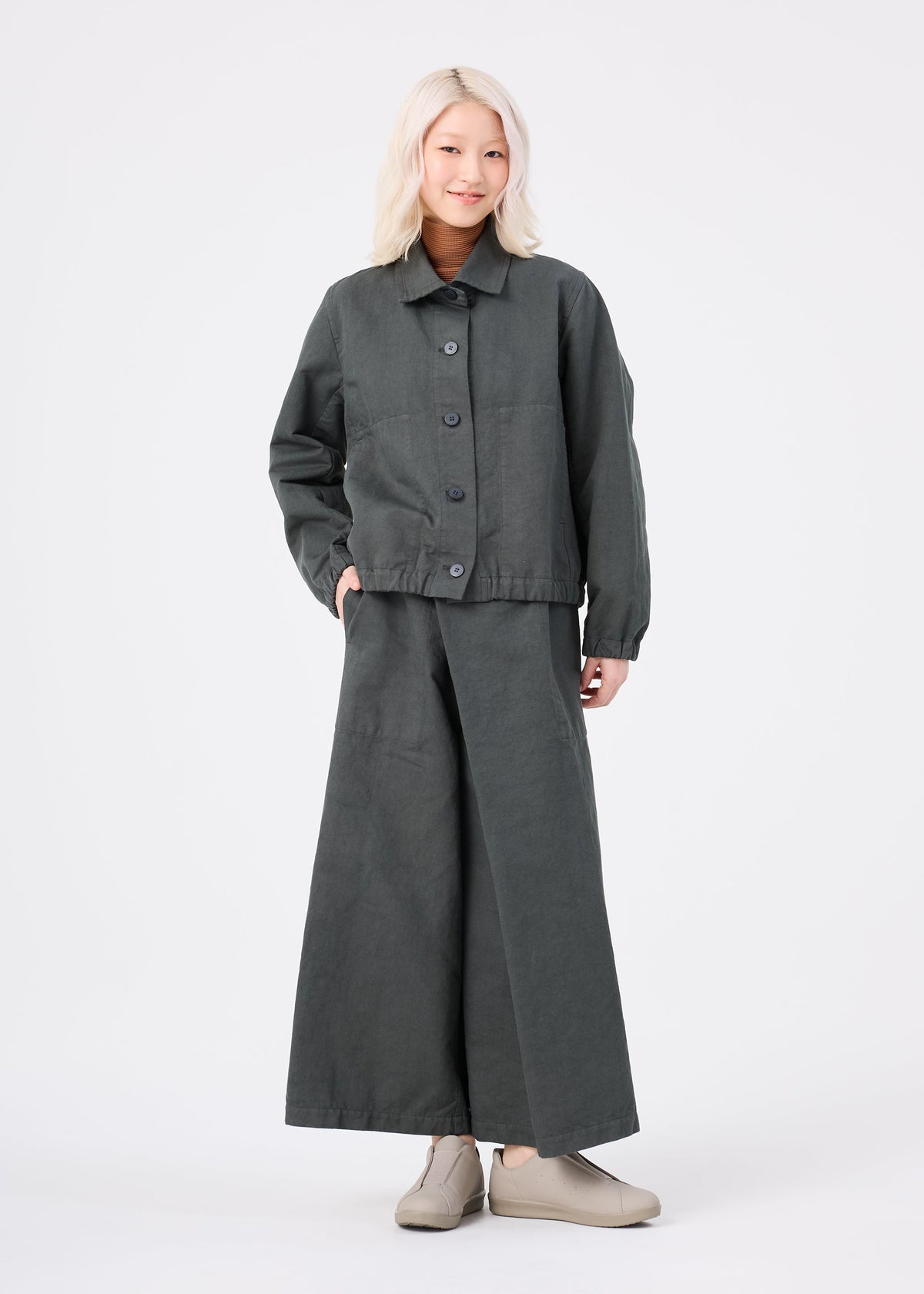 A person stands against a white background, wearing the dark gray PINA CLOTH jacket and wide-leg pants from me ISSEY MIYAKE with beige shoes. They have light blonde hair and are smiling slightly, with one hand in their jacket pocket. view 5 of 6