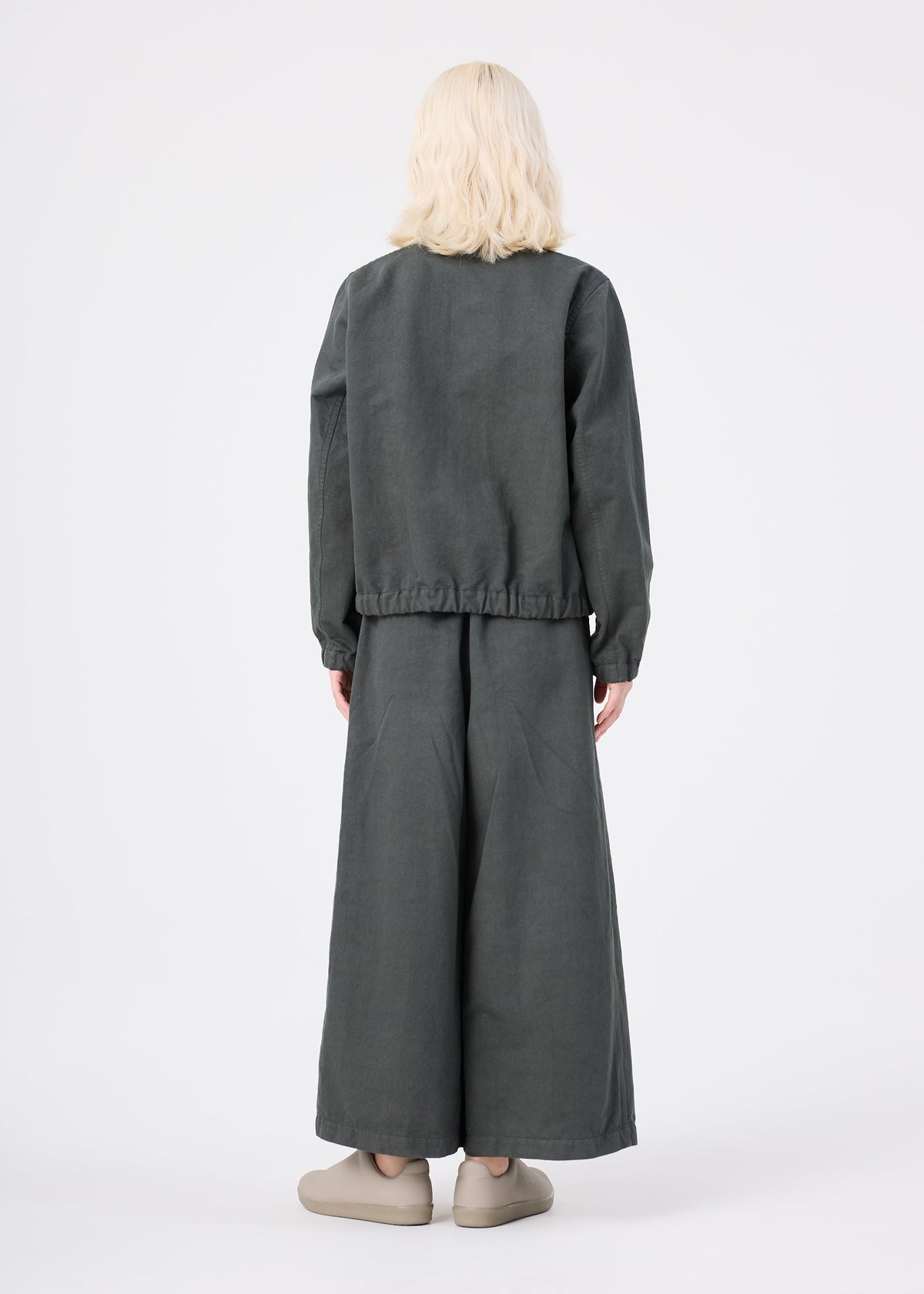 A person with light blonde hair stands facing away, dressed in a loose-fitting dark gray PINA CLOTH jacket and wide-legged matching pants by me ISSEY MIYAKE. They are wearing beige slip-on shoes, set against a plain white background. view 6 of 6