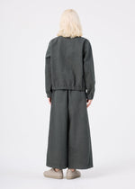 A person with light blonde hair stands facing away, dressed in a loose-fitting dark gray PINA CLOTH jacket and wide-legged matching pants by me ISSEY MIYAKE. They are wearing beige slip-on shoes, set against a plain white background.