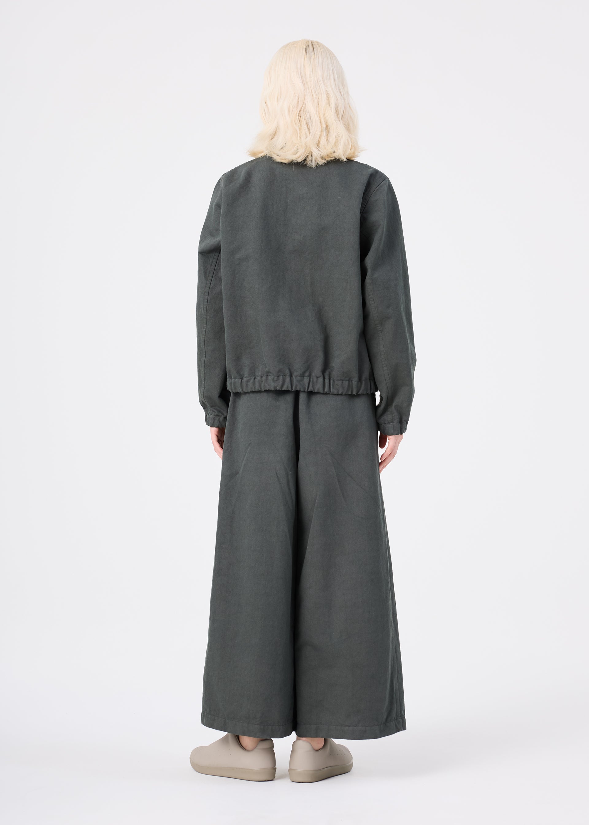 Zoomed view A person with light blonde hair stands facing away, dressed in a loose-fitting dark gray PINA CLOTH jacket and wide-legged matching pants by me ISSEY MIYAKE. They are wearing beige slip-on shoes, set against a plain white background.