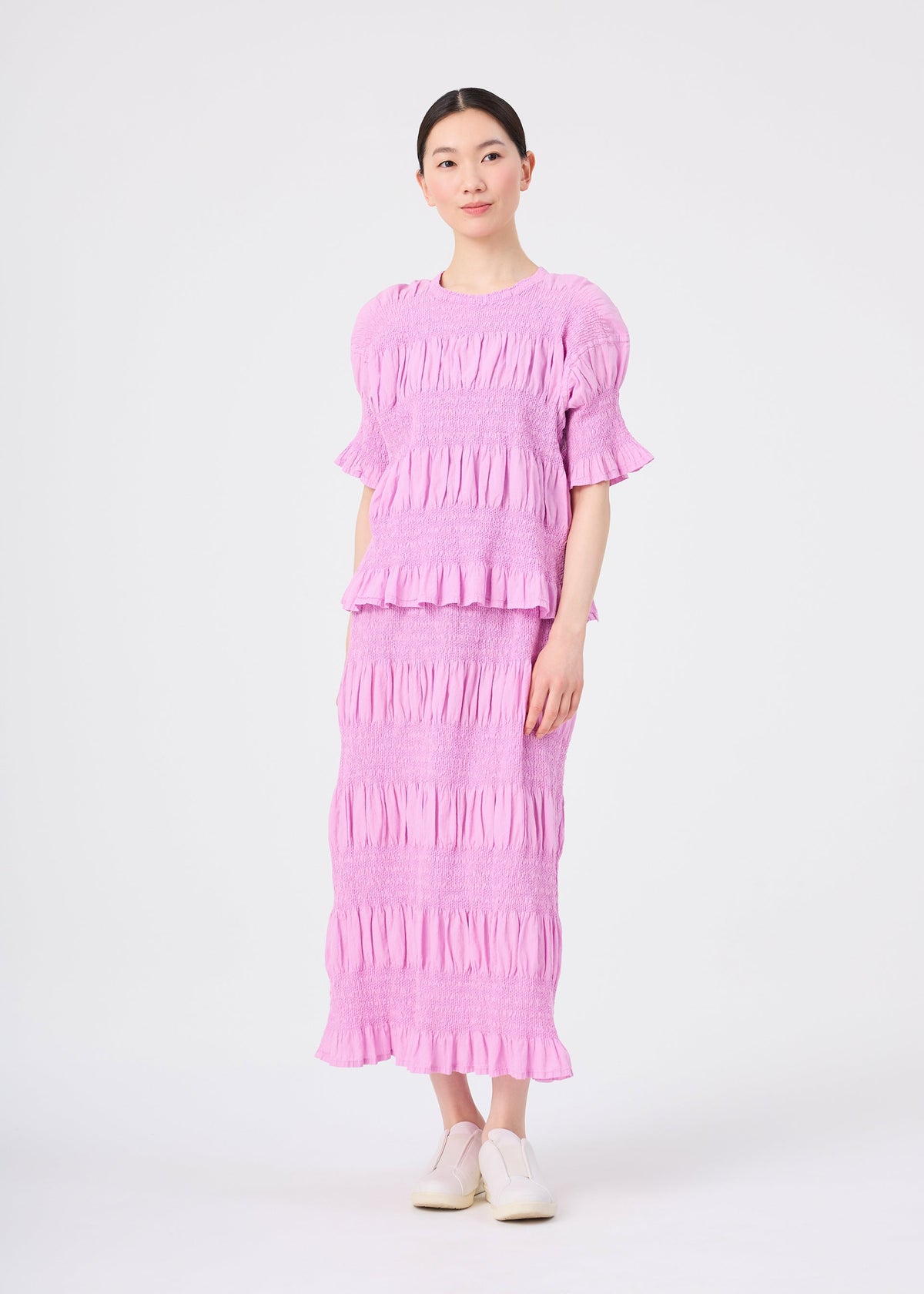 A person stands against a plain background, wearing the me ISSEY MIYAKE SHRINK STRIPE set—a matching light purple top and a cotton blend skirt featuring ruffled details and crafted with a unique product-dyeing technique. The look is completed with white shoes. view 5 of 6