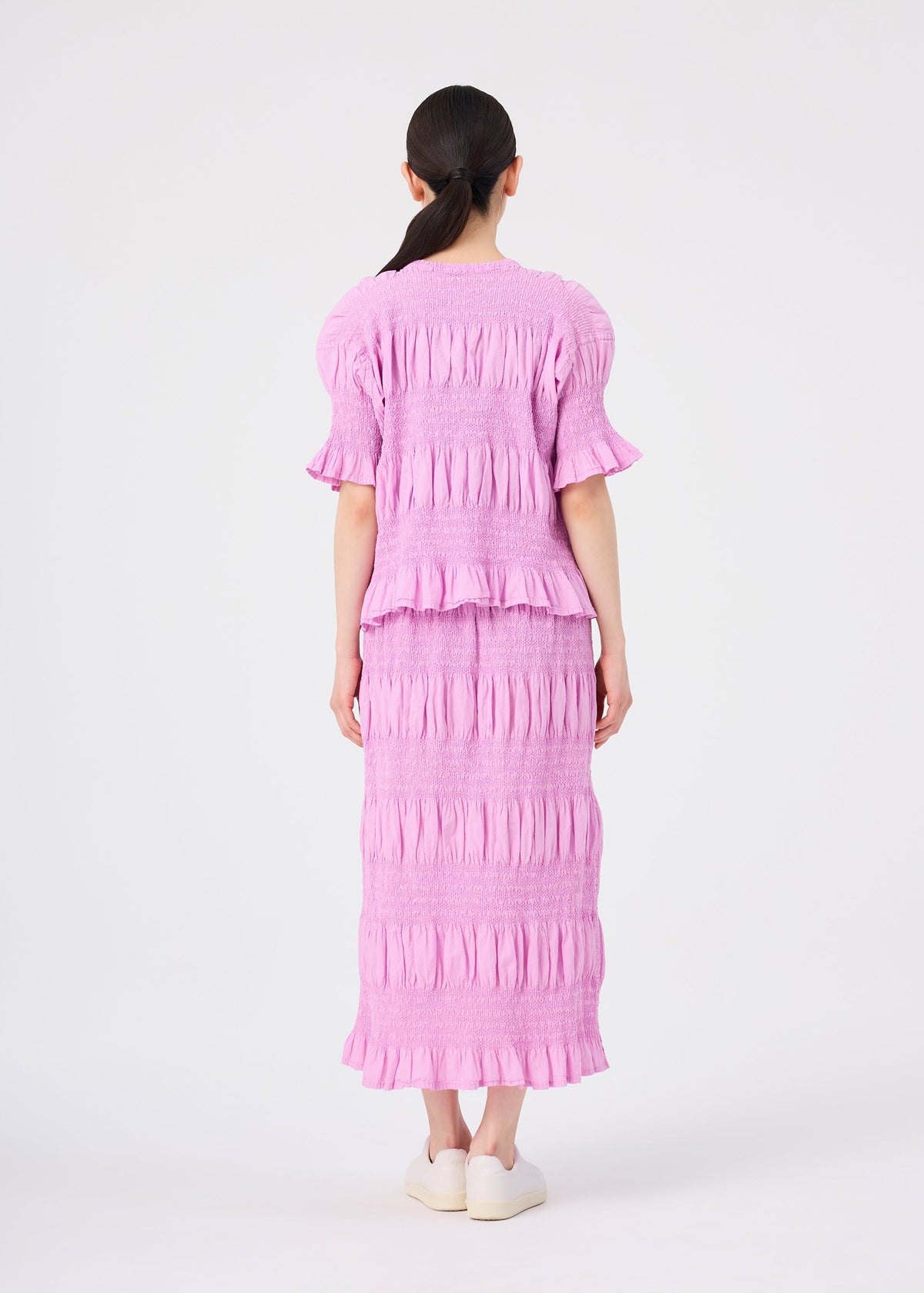 A person with long dark hair, wearing the me ISSEY MIYAKE SHRINK STRIPE matching light purple textured top and cotton blend skirt with ruffle details—crafted using a special product-dyeing technique—stands facing away against a plain white background. White slip-on shoes complete the outfit. view 6 of 6