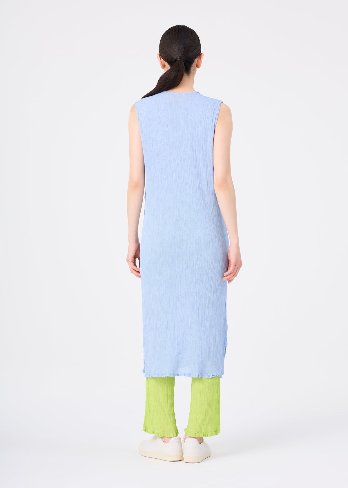A person with long dark hair, wearing the light blue ME ISSEY MIYAKE SEE-THROUGH CREPE COLOR sleeveless dress over bright green pleated pants and white shoes, stands facing away against a plain white background. view 7 of 8