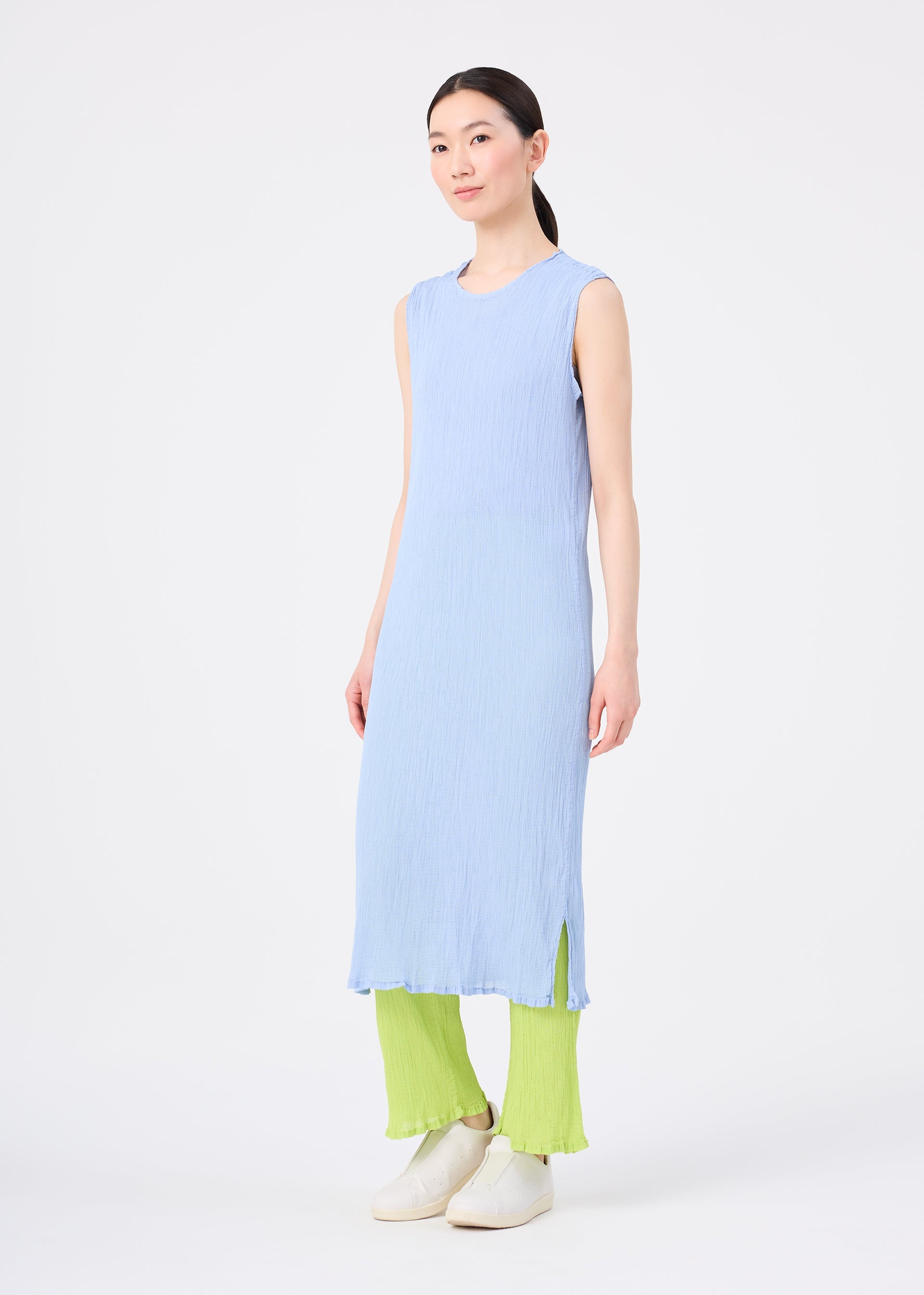 Zoomed view A person stands against a plain white background wearing the sleeveless SEE-THROUGH CREPE COLOR dress by me ISSEY MIYAKE, paired with lime green pleated pants and white slip-on shoes.