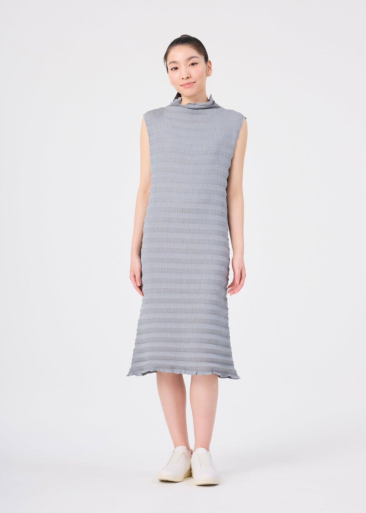 A young East Asian woman stands facing forward, dressed in the ACCORDION SEE-THROUGH CREPE midi dress from me ISSEY MIYAKE—a sleeveless, light gray piece featuring garment-dyed fabric, delicate accordion hand pleating, and a high neckline. She pairs the dress with matching slip-on shoes against a plain light gray background. view 5 of 6