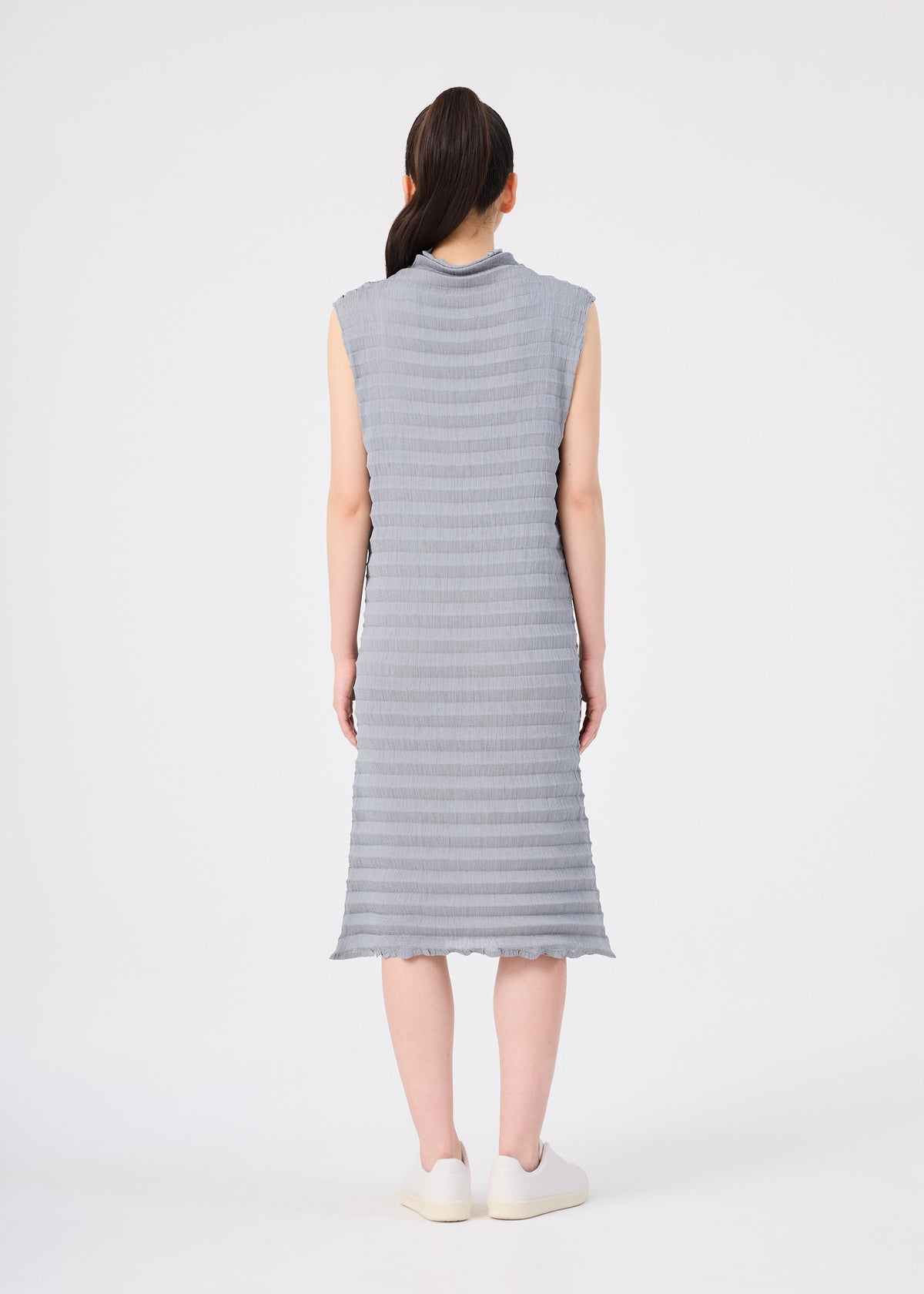 A person with long dark hair in a ponytail is seen from behind, wearing the me ISSEY MIYAKE ACCORDION SEE-THROUGH CREPE sleeveless, knee-length textured gray dress made from garment-dyed fabric, paired with white shoes, standing against a plain white background. view 6 of 6