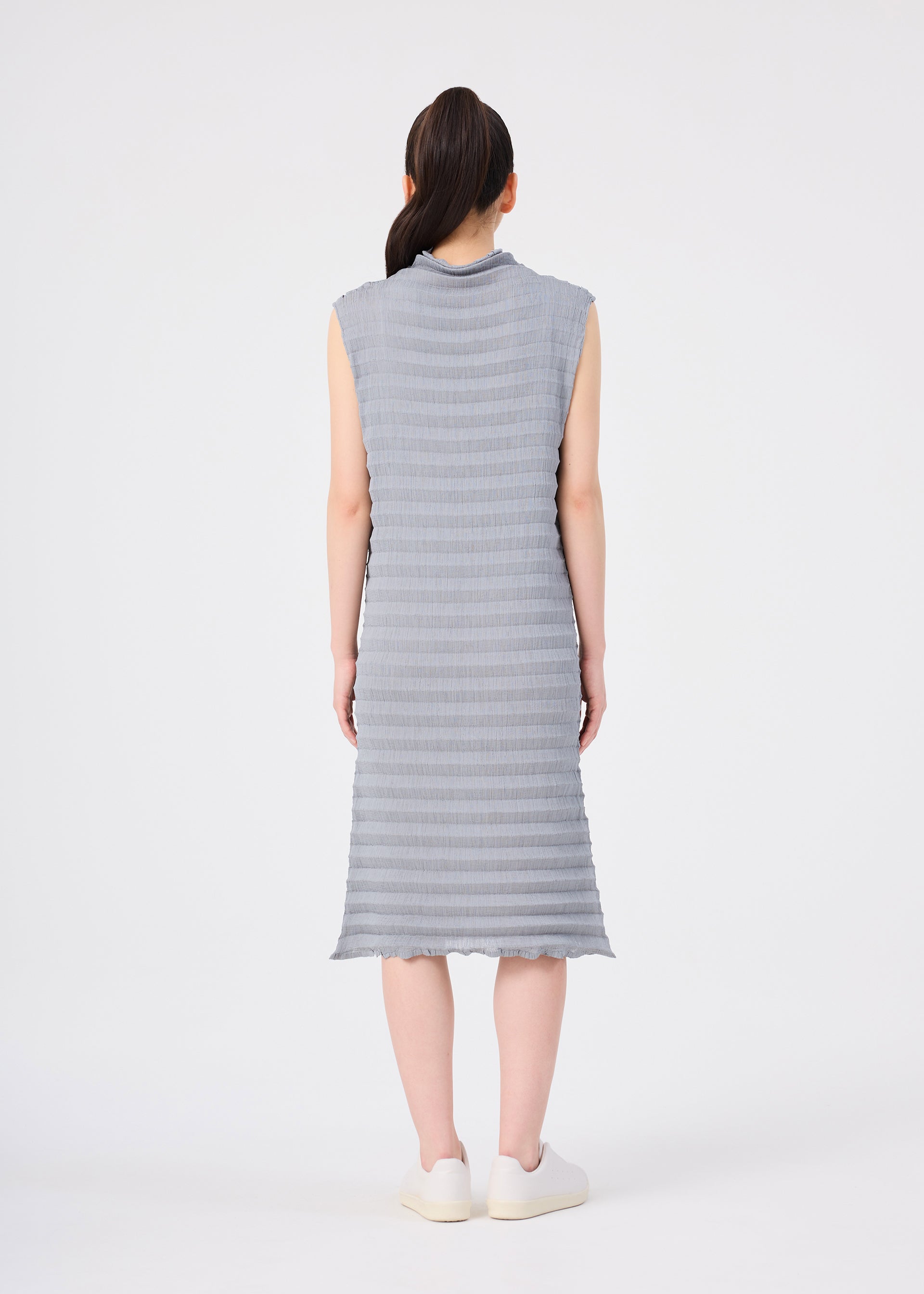 Zoomed view A person with long dark hair in a ponytail is seen from behind, wearing the me ISSEY MIYAKE ACCORDION SEE-THROUGH CREPE sleeveless, knee-length textured gray dress made from garment-dyed fabric, paired with white shoes, standing against a plain white background.