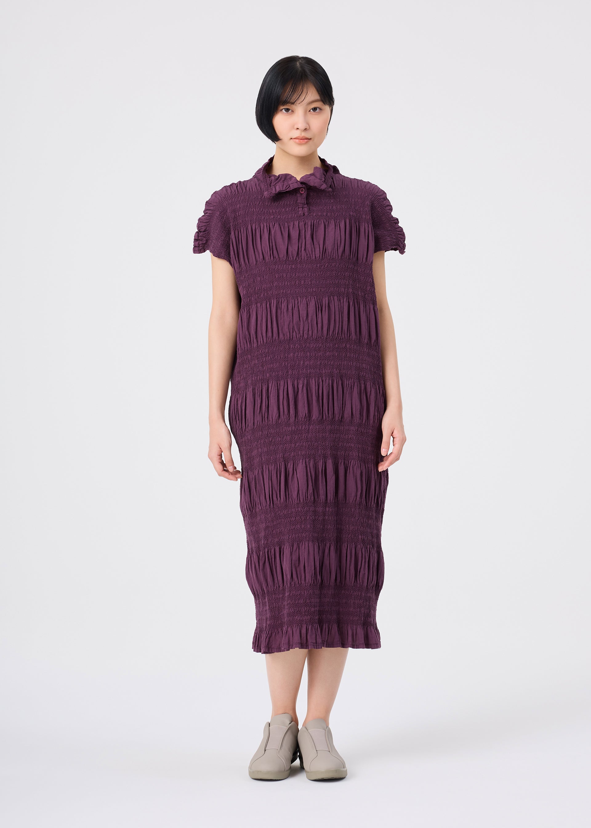 Zoomed view A person with short dark hair wears the SHRINK STRIPE knee-length cotton-blend dress by me ISSEY MIYAKE in a textured purple design with short sleeves, paired with light-colored shoes, standing against a plain white background.