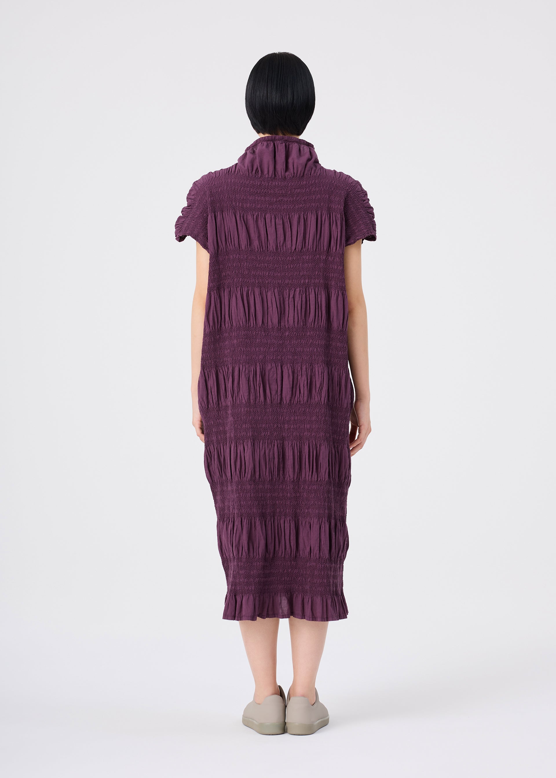 Zoomed view A person with short black hair stands facing away, wearing the SHRINK STRIPE knee-length dress by me ISSEY MIYAKE—a textured, purple collared dress with short sleeves and a high collar—crafted from a soft cotton-blend fabric. The look is completed with light-colored shoes and set against a plain white background.