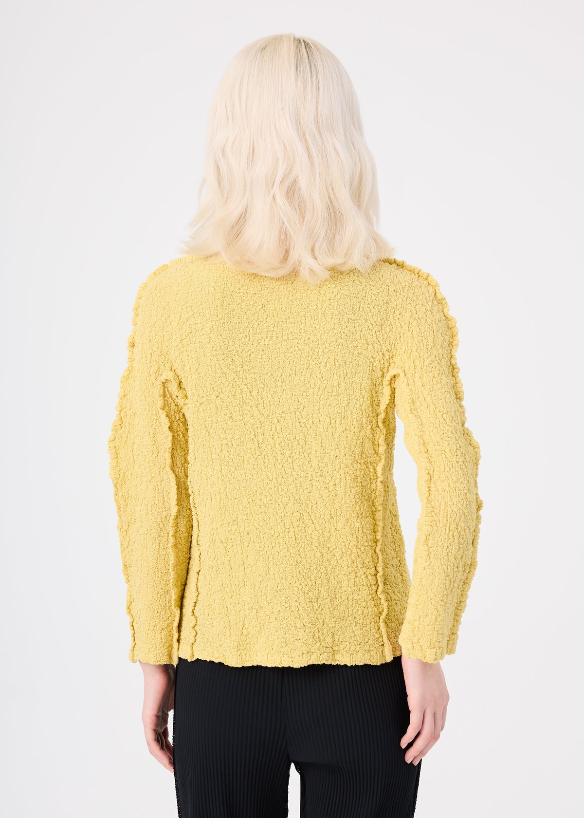 Zoomed view A person with shoulder-length, wavy blonde hair faces away, wearing the me ISSEY MIYAKE ROUND CAULIFLOWER yellow textured long-sleeve top and black pants against a plain white background.