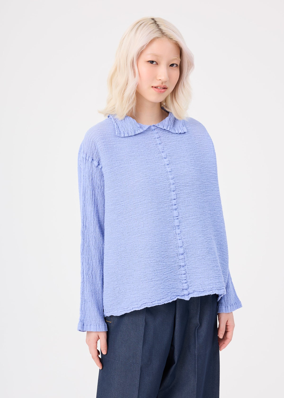 A person with wavy, light blonde hair wears the me ISSEY MIYAKE POLYCOTTON CREPE: a textured, light blue collared top and wide navy cotton-polyester pants, standing against a plain white background. view 6 of 8