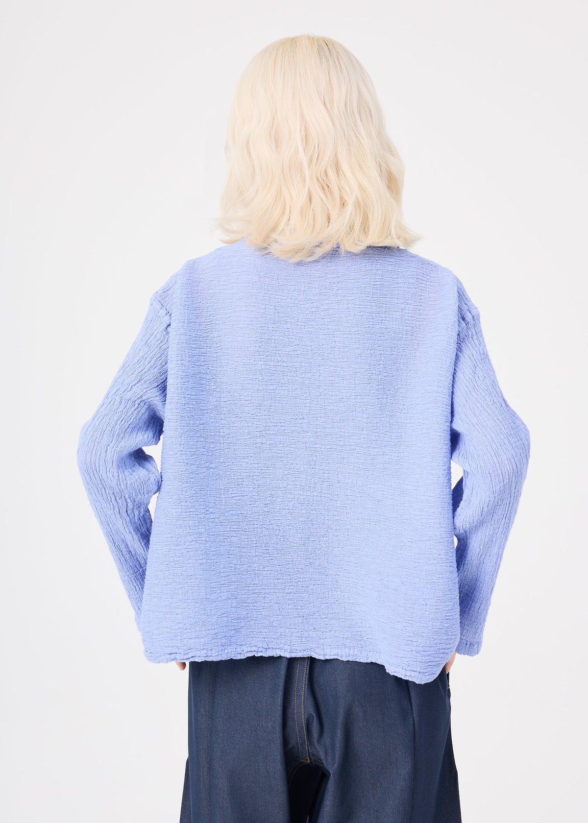 A person with wavy blonde hair, wearing the me ISSEY MIYAKE POLYCOTTON CREPE textured light blue long-sleeved top and dark pants, stands with their back to the camera against a white background. view 7 of 8