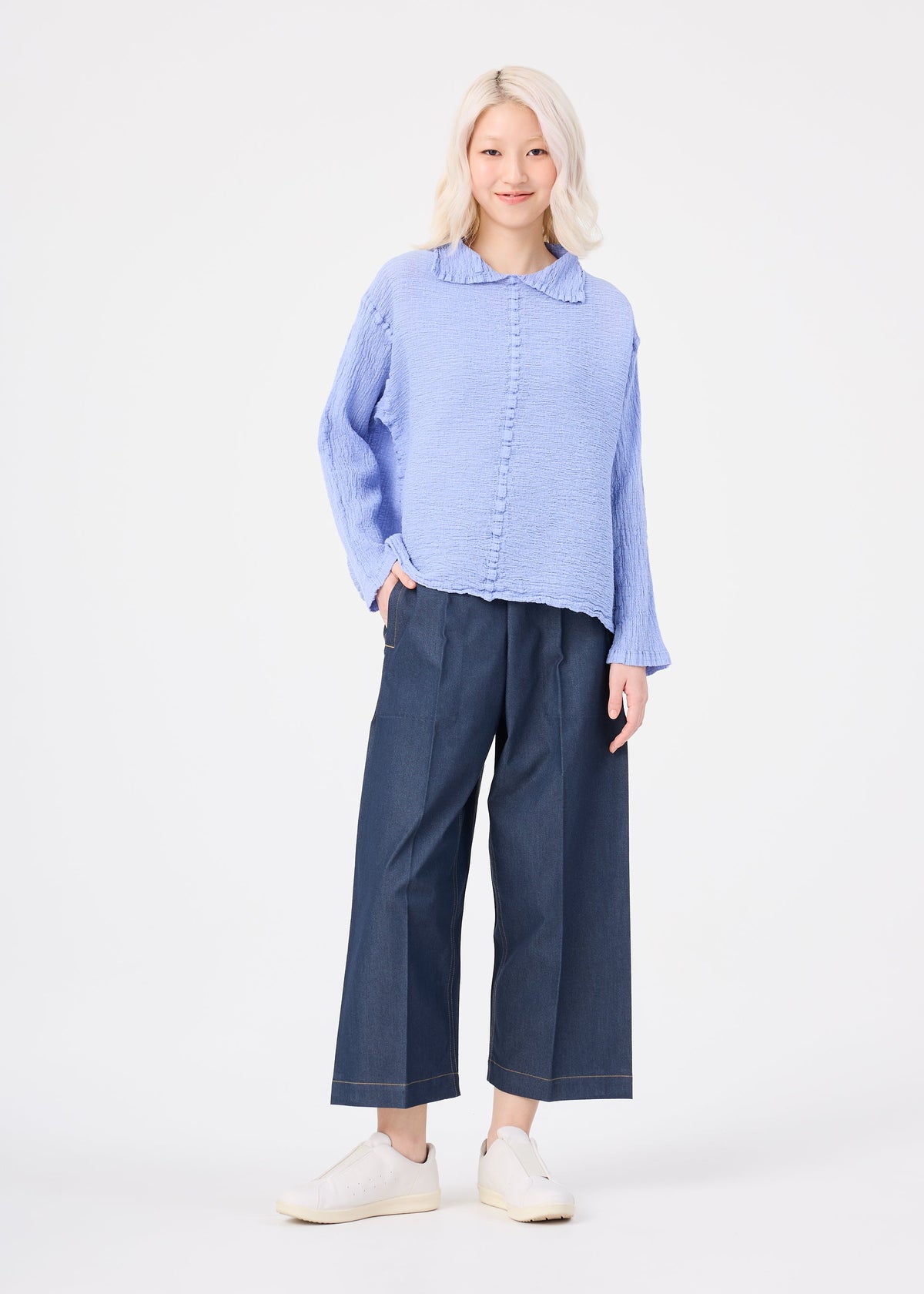 Zoomed view A person with short, light blonde hair wears the POLYCOTTON CREPE long-sleeved top by me ISSEY MIYAKE, wide dark blue pants, and white shoes, smiling against a plain white background.