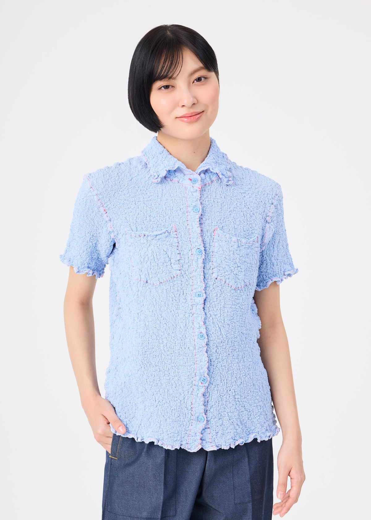 A person with short black hair wears the me ISSEY MIYAKE CAULIFLOWER COLOR 1 light blue textured cotton short-sleeved collared top with two chest pockets and dark blue pants, standing against a plain white background and slightly smiling. view 5 of 7