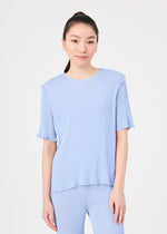 A woman with long dark hair in a ponytail wears the SEE-THROUGH CREPE COLOR set by me ISSEY MIYAKE—light blue, short-sleeved top and matching pants—standing against a white background and smiling gently.