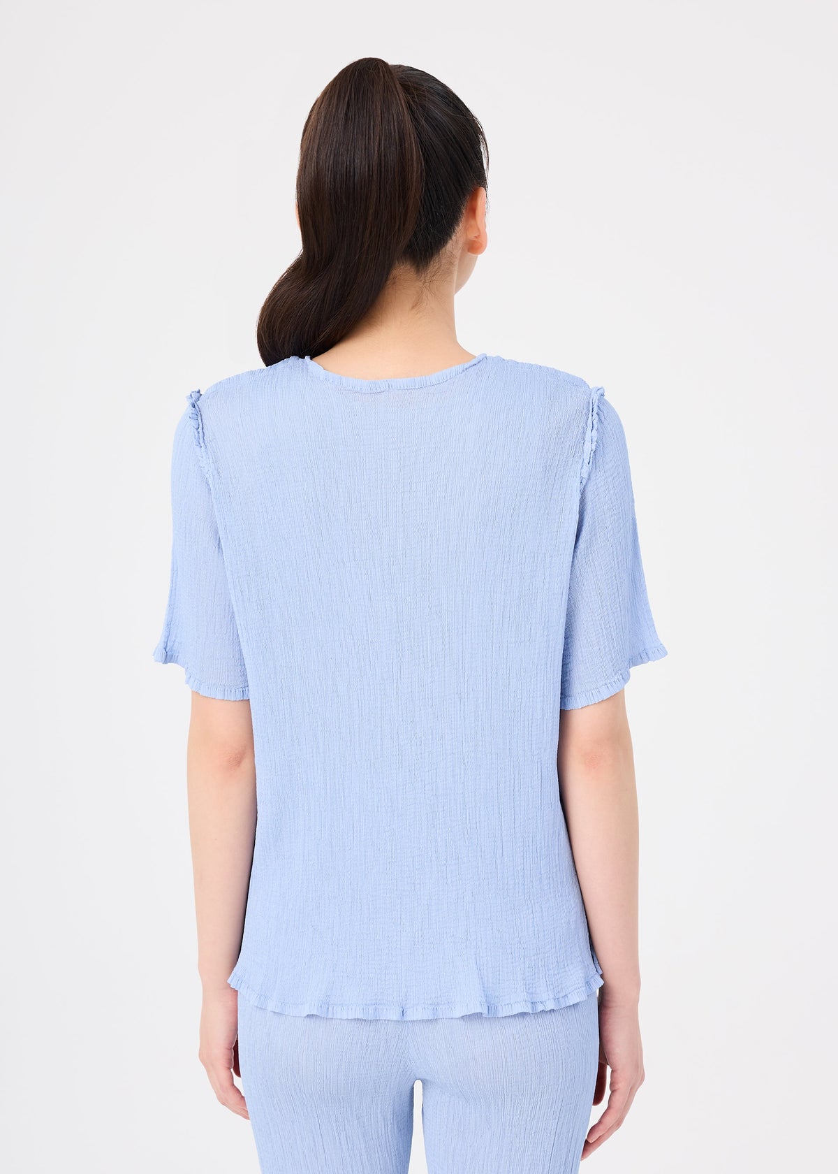 A woman with long dark hair in a ponytail is shown from behind, wearing the SEE-THROUGH CREPE COLOR set by me ISSEY MIYAKE—light blue, product-dyed sheer top and matching pants—against a plain white background. view 6 of 7