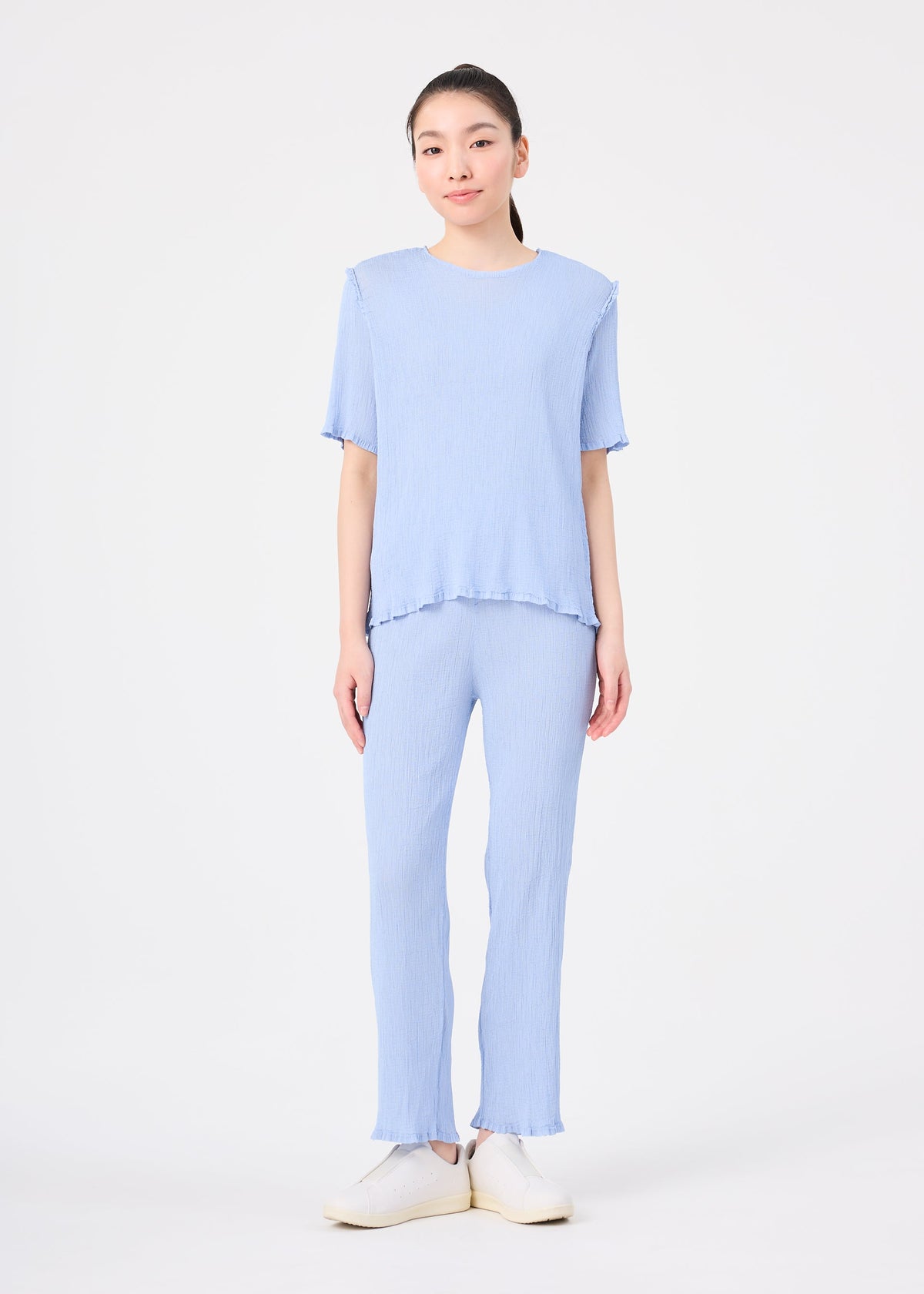 A person stands facing forward in the me ISSEY MIYAKE SEE-THROUGH CREPE COLOR light blue short-sleeve top and matching relaxed-fit pants, paired with white slip-on shoes, against a plain white background. view 7 of 7