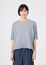 A person with short black hair wears the me ISSEY MIYAKE ACCORDION SEE-THROUGH CREPE, a loose, textured gray garment-dyed top, paired with navy pants and standing against a plain white background.