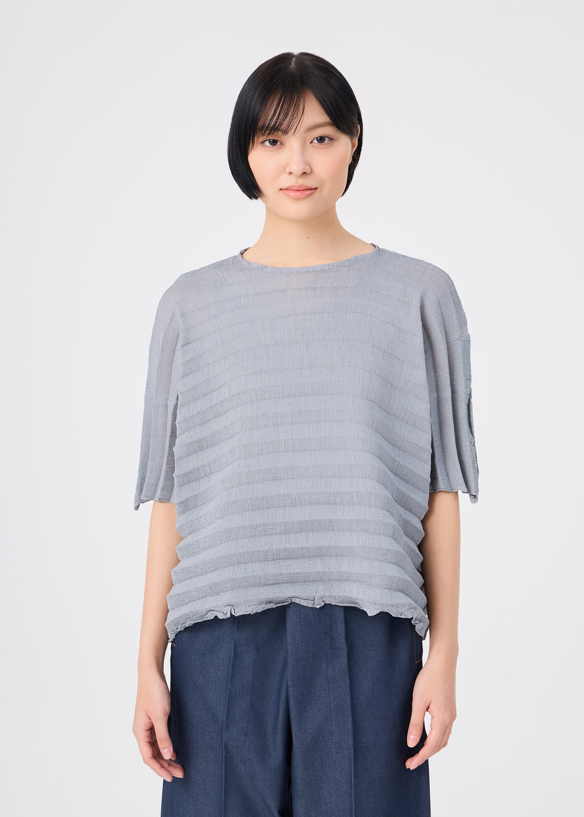Zoomed view A person with short black hair wears the me ISSEY MIYAKE ACCORDION SEE-THROUGH CREPE, a loose, textured gray garment-dyed top, paired with navy pants and standing against a plain white background.