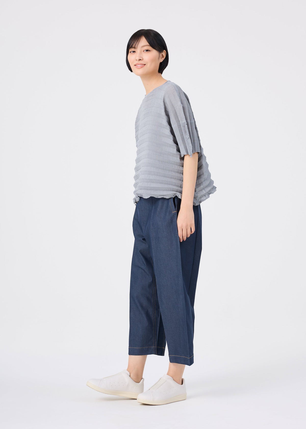 A person stands against a white background wearing the ACCORDION SEE-THROUGH CREPE top from me ISSEY MIYAKE, featuring textured, garment-dyed gray fabric. They pair it with loose dark blue pants and white sneakers, looking slightly to the side and smiling. view 7 of 7