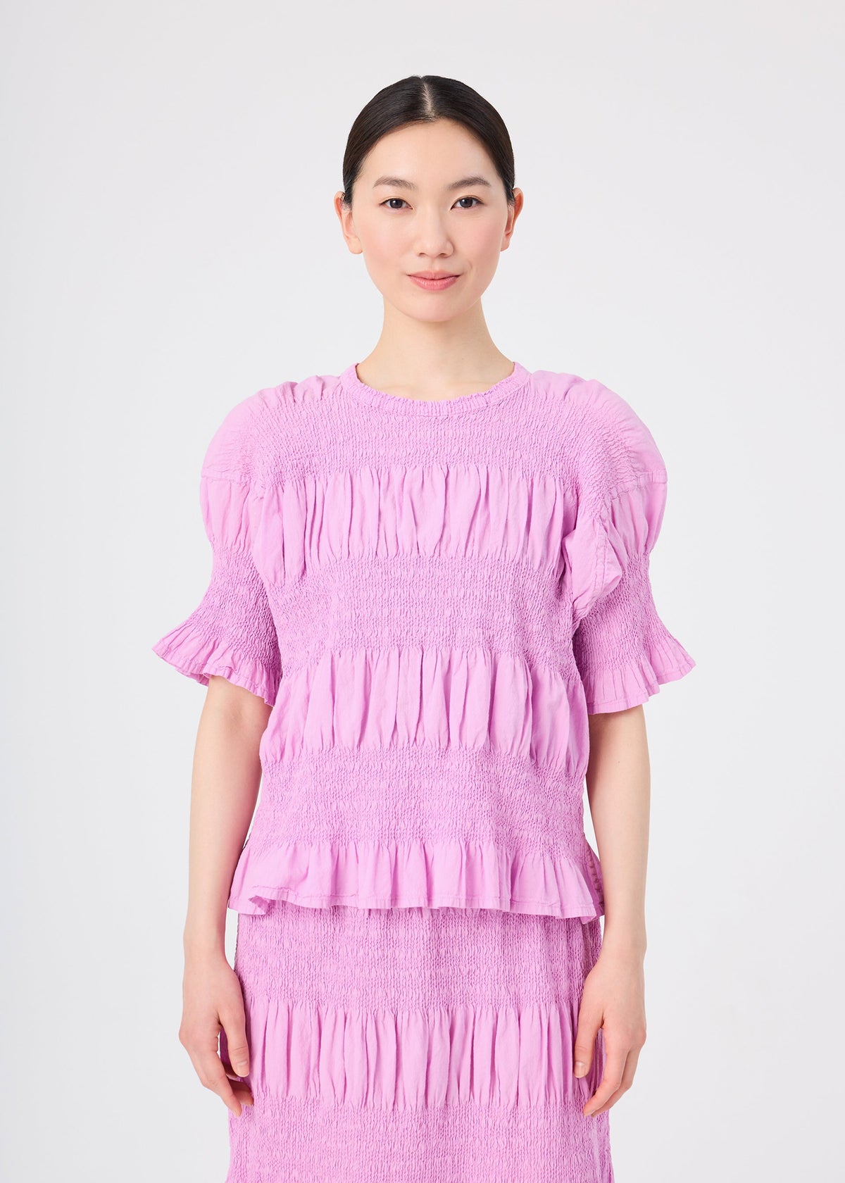 A woman with dark hair tied back is wearing the me ISSEY MIYAKE SHRINK STRIPE lightweight, light purple, short-sleeve ruffled blouse and matching skirt. She is standing and smiling against a plain white background. view 5 of 7