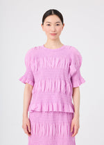 A woman with dark hair tied back is wearing the me ISSEY MIYAKE SHRINK STRIPE lightweight, light purple, short-sleeve ruffled blouse and matching skirt. She is standing and smiling against a plain white background.