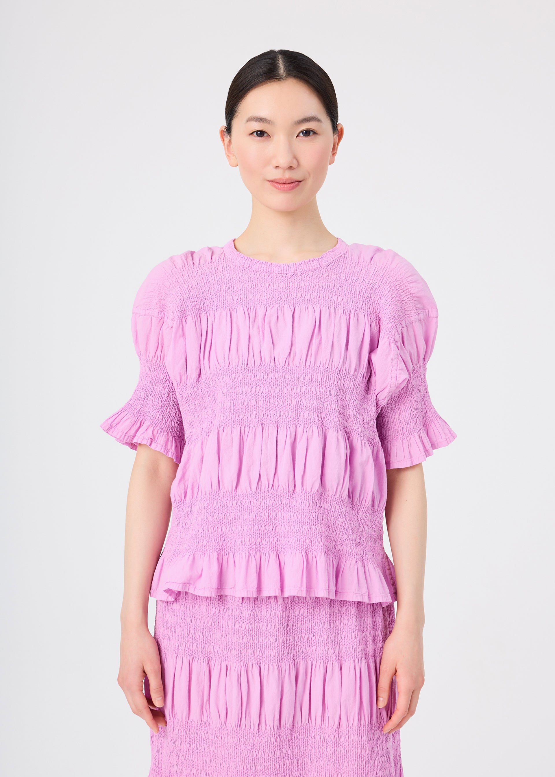 Zoomed view A woman with dark hair tied back is wearing the me ISSEY MIYAKE SHRINK STRIPE lightweight, light purple, short-sleeve ruffled blouse and matching skirt. She is standing and smiling against a plain white background.