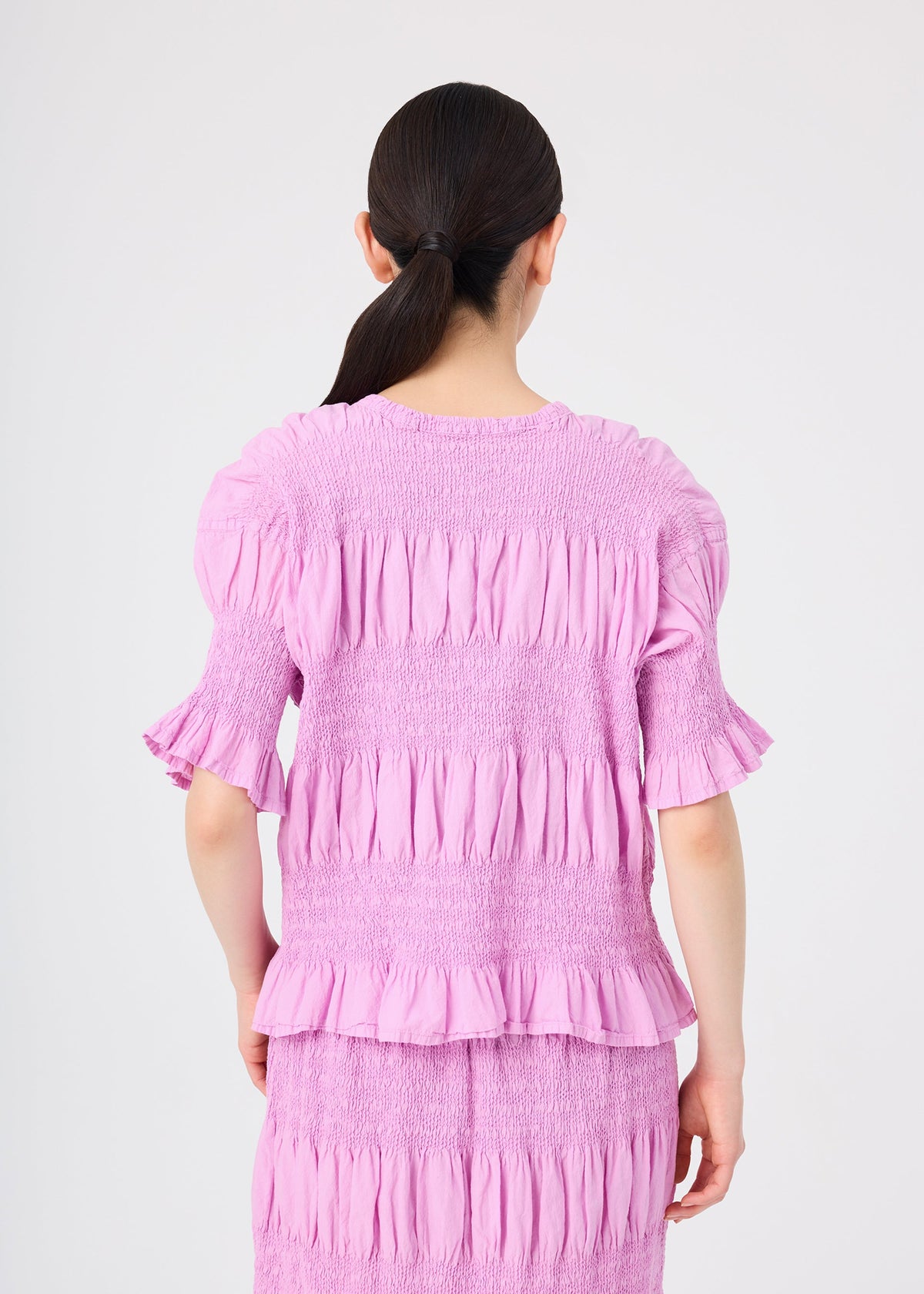 A woman with dark hair in a low ponytail is shown from behind, wearing the me ISSEY MIYAKE SHRINK STRIPE lightweight short-sleeve top and matching skirt in light purple against a plain light background. view 6 of 7