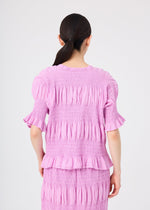 A woman with dark hair in a low ponytail is shown from behind, wearing the me ISSEY MIYAKE SHRINK STRIPE lightweight short-sleeve top and matching skirt in light purple against a plain light background.