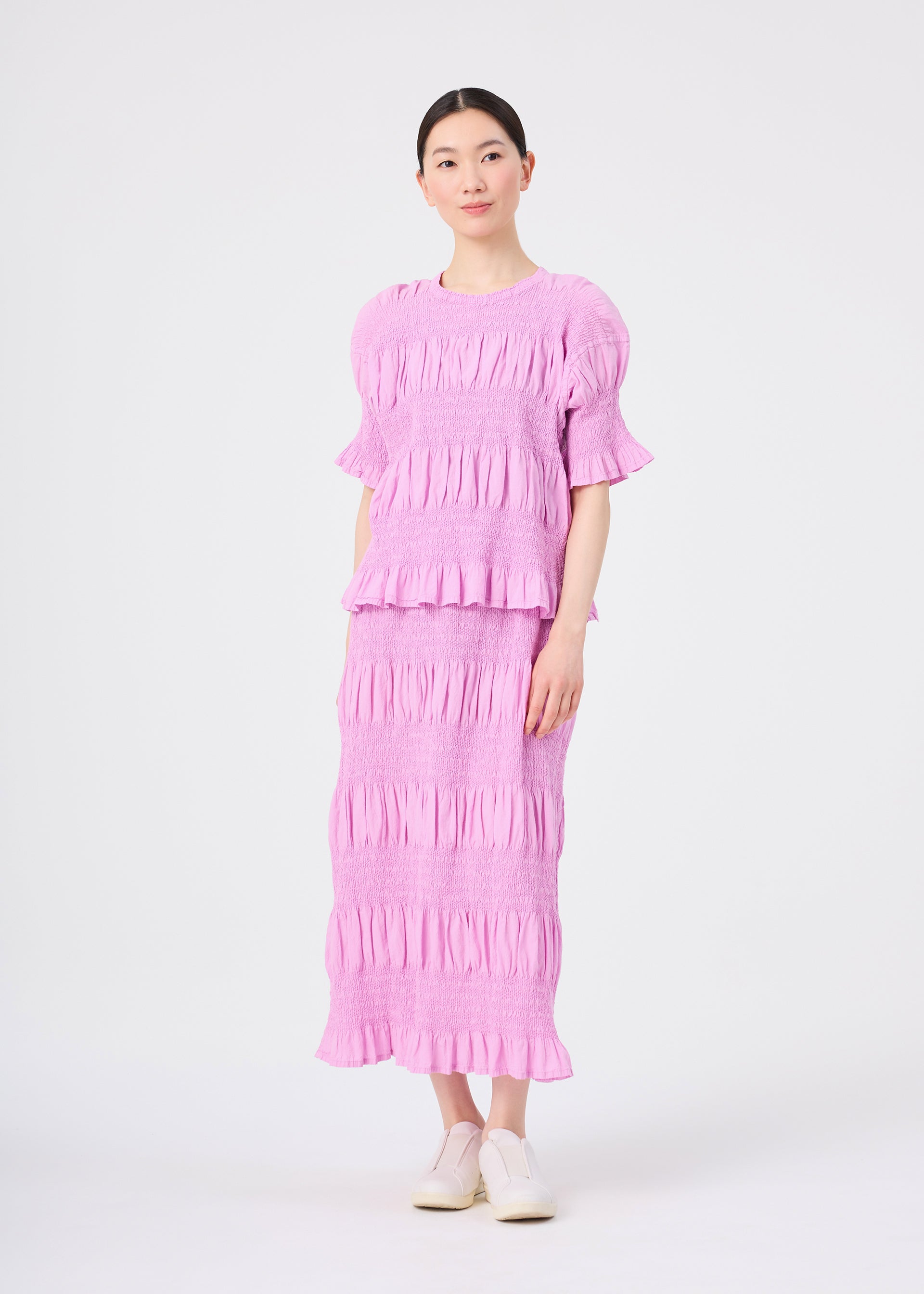 Zoomed view A woman stands against a white background wearing the me ISSEY MIYAKE SHRINK STRIPE lightweight, light purple ruffled top and matching skirt, paired with white shoes.