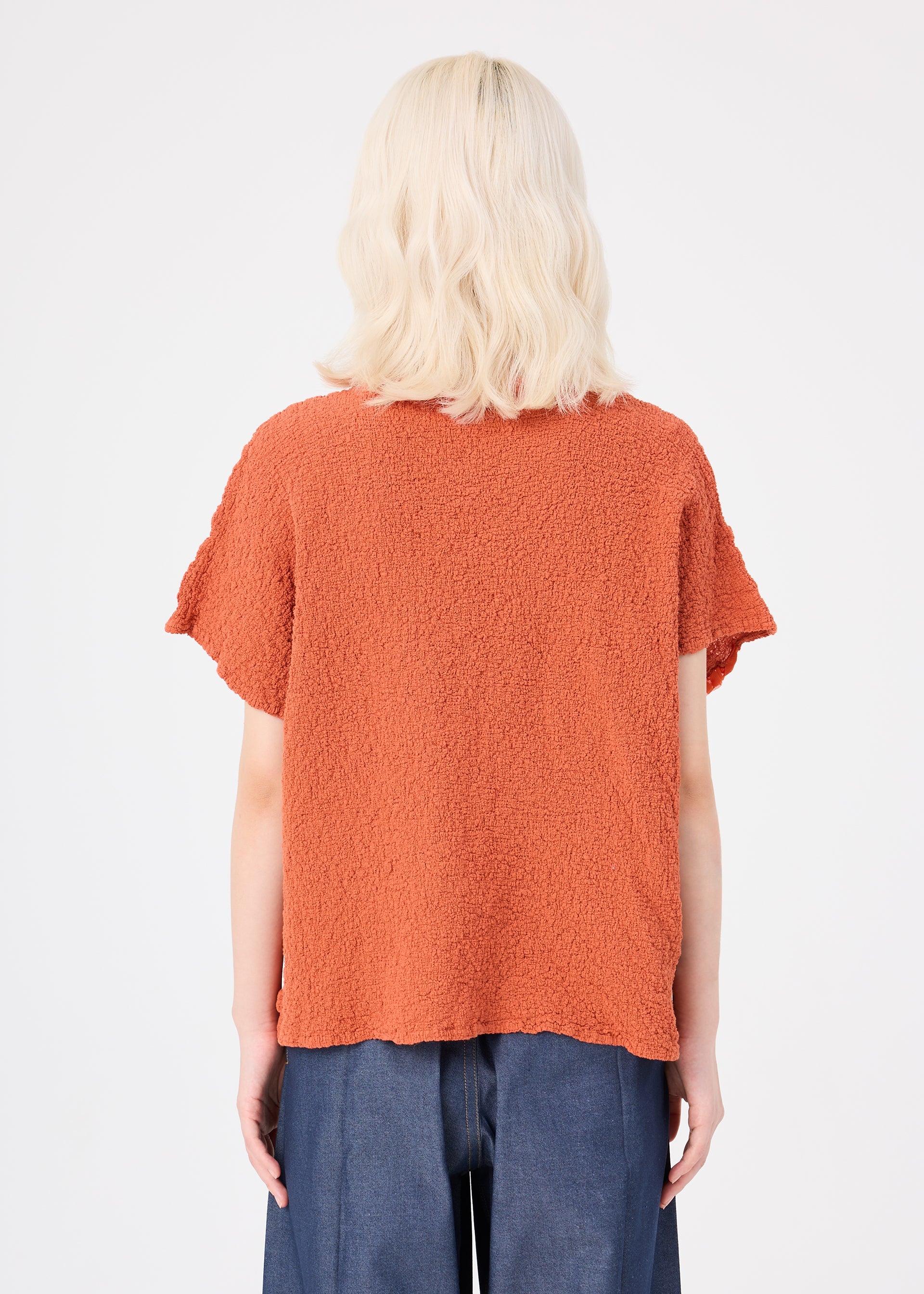 Zoomed view A person with shoulder-length blonde hair is seen from behind, standing against a plain white background and wearing the me ISSEY MIYAKE LEAF TULLE CAULIFLOWER top—an orange cotton short-sleeved piece with a relaxed square silhouette—paired with blue jeans.