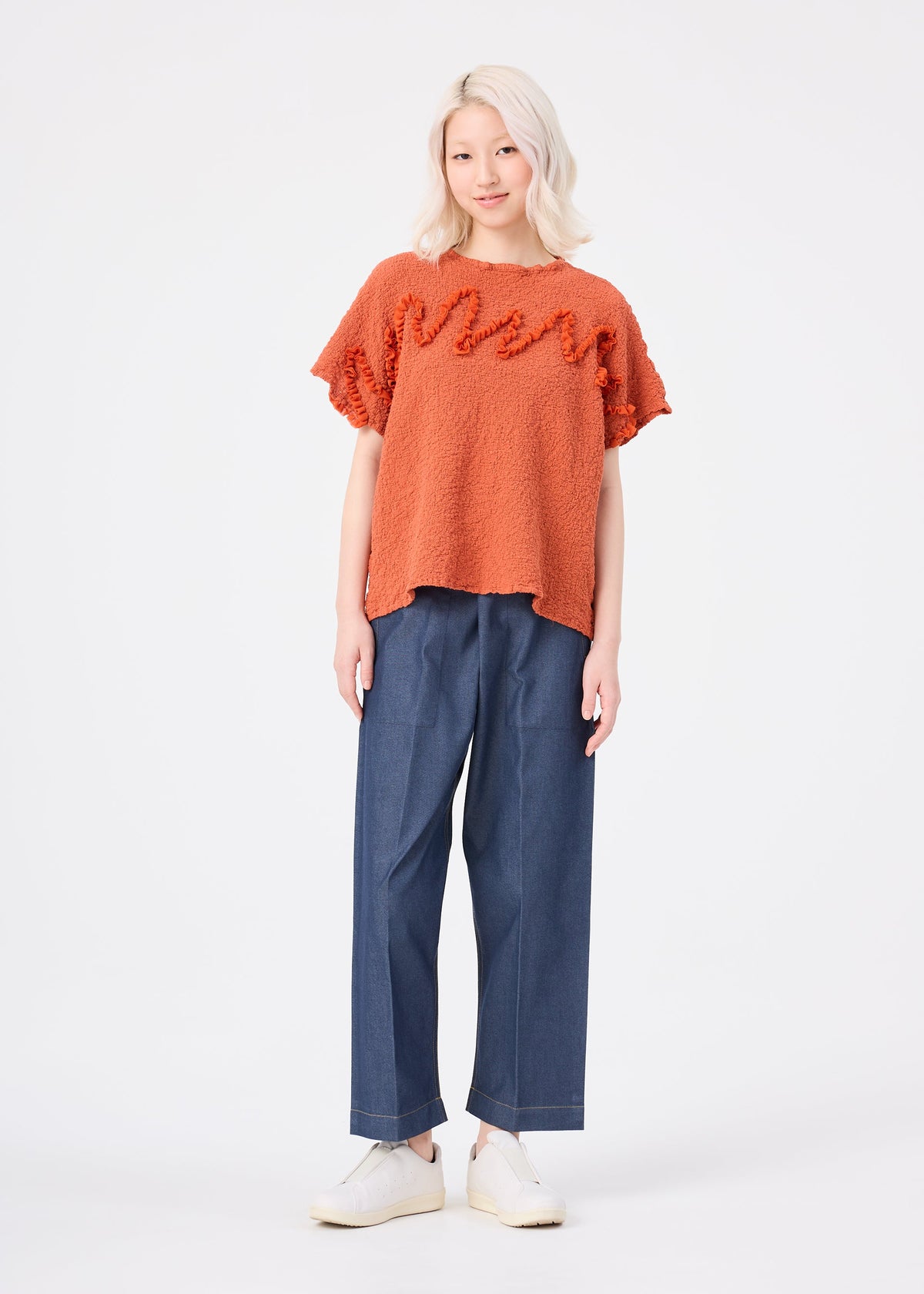 A person with light hair wearing the LEAF TULLE CAULIFLOWER textured cotton short-sleeved top by me ISSEY MIYAKE in orange, paired with wide-leg blue jeans and white shoes, stands facing forward in a studio against a plain white background. The relaxed square silhouette of the top from me ISSEY MIYAKE adds a modern touch to the look. view 7 of 7
