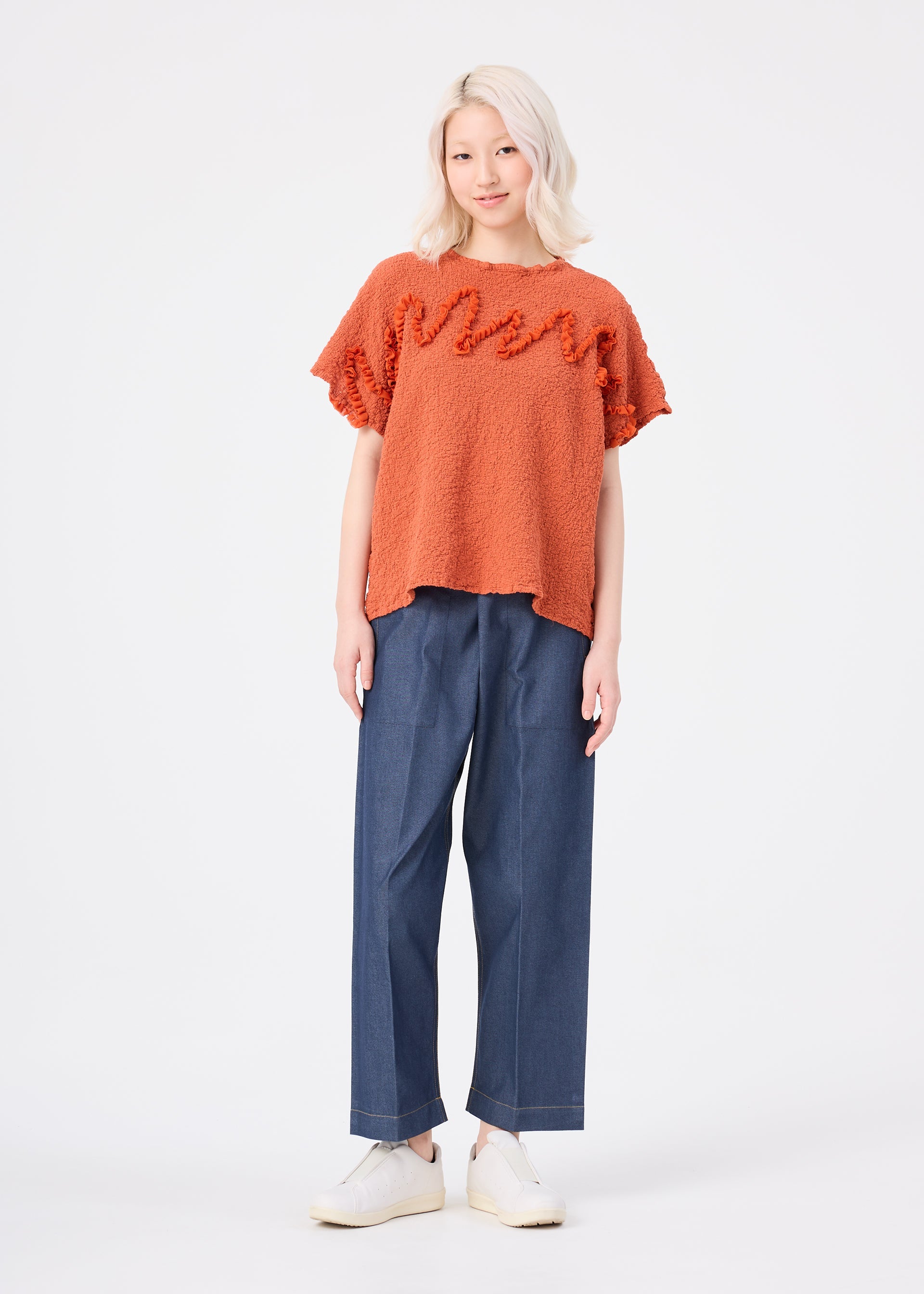 Zoomed view A person with light hair wearing the LEAF TULLE CAULIFLOWER textured cotton short-sleeved top by me ISSEY MIYAKE in orange, paired with wide-leg blue jeans and white shoes, stands facing forward in a studio against a plain white background. The relaxed square silhouette of the top from me ISSEY MIYAKE adds a modern touch to the look.