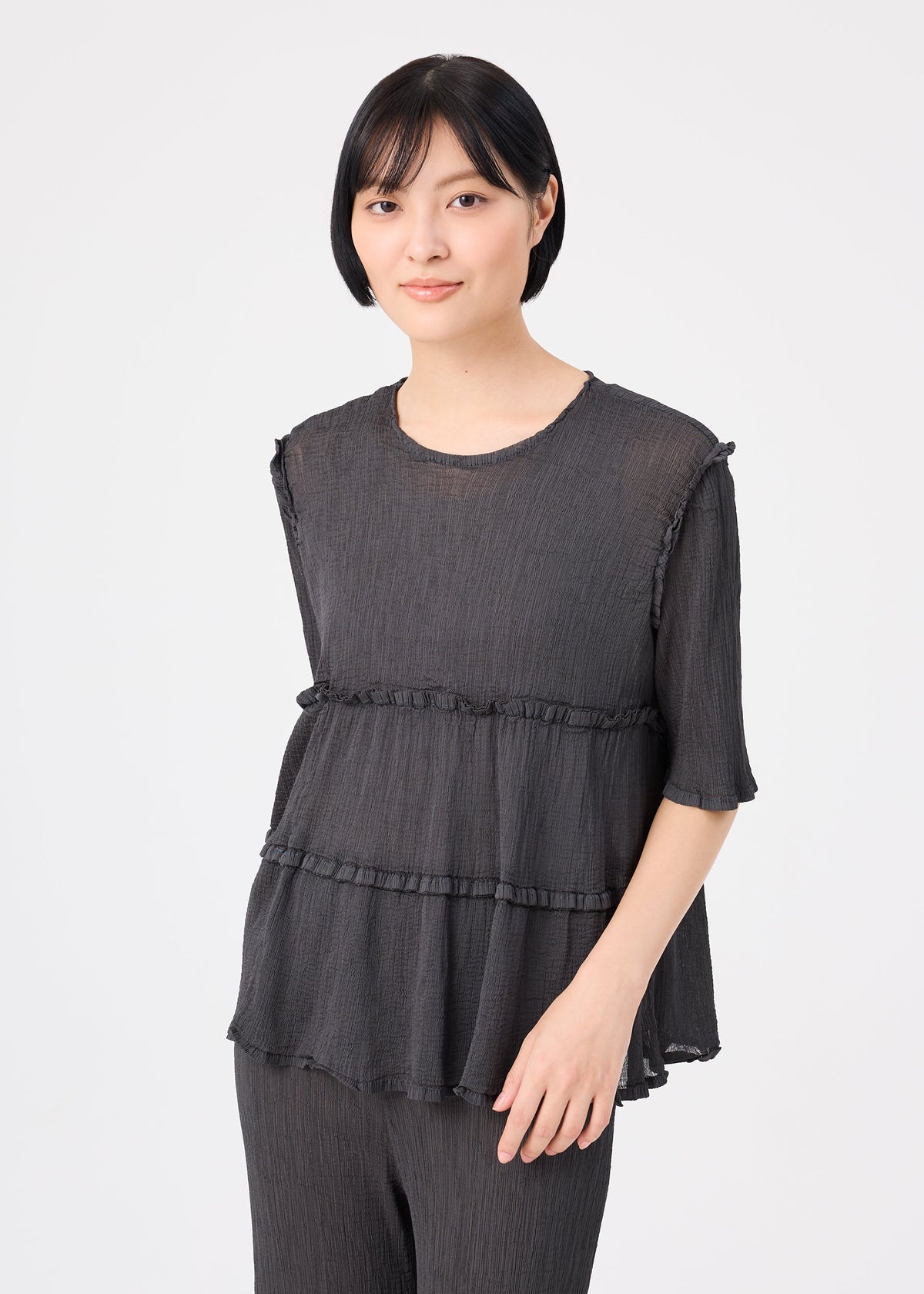 A person with short black hair wears the FLARE SEE-THROUGH CREPE set by me ISSEY MIYAKE—featuring a loose, dark, short-sleeved crepe shirt with tiered detailing and matching pants—standing against a plain light background and looking at the camera with a slight smile. view 5 of 7