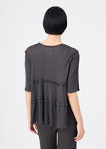 A person with short black hair is seen from behind, standing against a plain light background and wearing the me ISSEY MIYAKE FLARE SEE-THROUGH CREPE set—loose, textured dark gray upcycled natural dye top with tiered ruffles and matching pants.