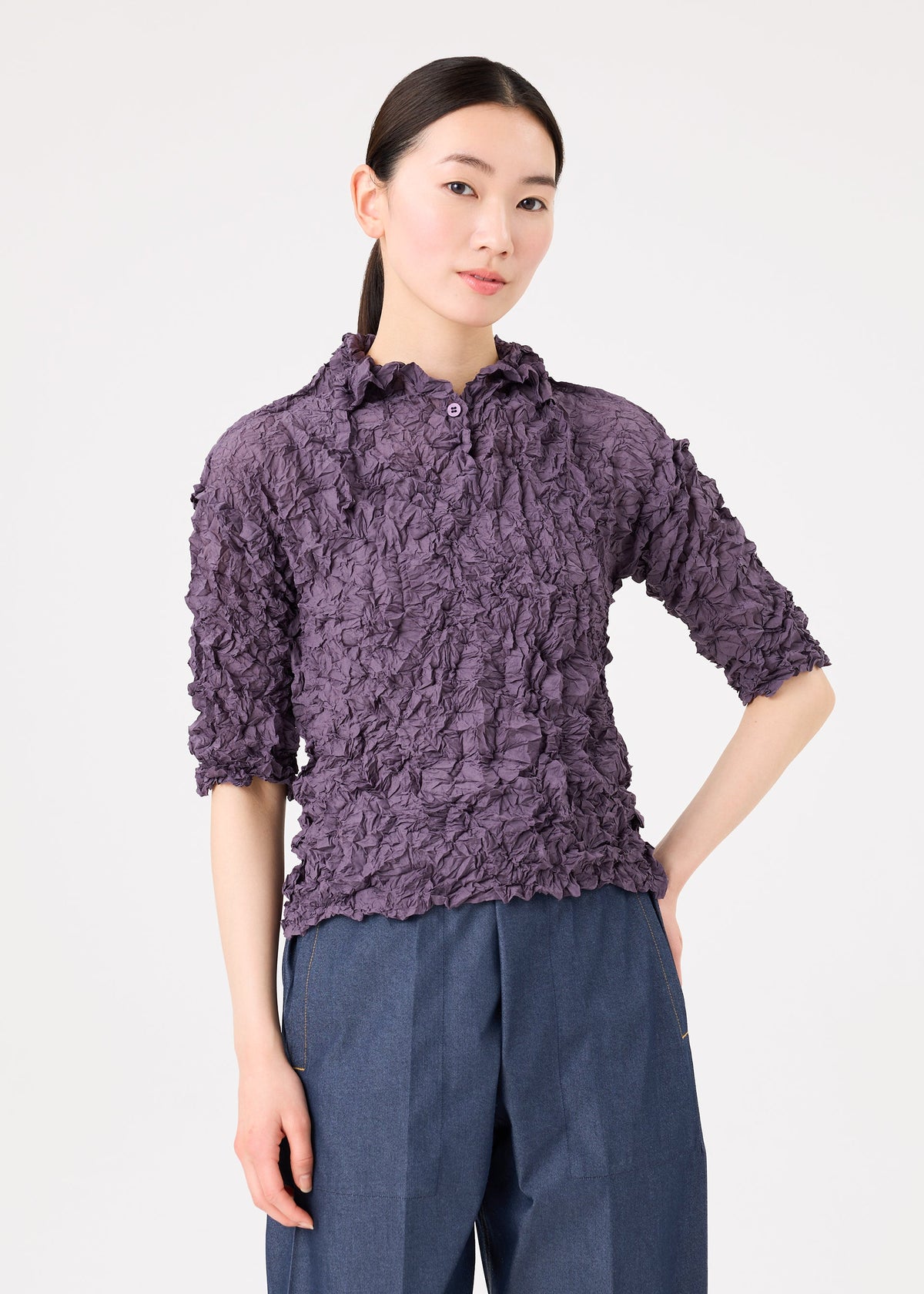 A woman poses against a white background, wearing the MERINGUE TOP by me ISSEY MIYAKE—a textured purple crushed pleat top with elbow-length sleeves—and blue pants, with one hand in her pocket. view 7 of 9