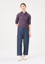A person stands against a white background wearing the MERINGUE TOP by me ISSEY MIYAKE in purple, paired with wide-leg blue pants and white shoes, hands behind their back and a slight smile.