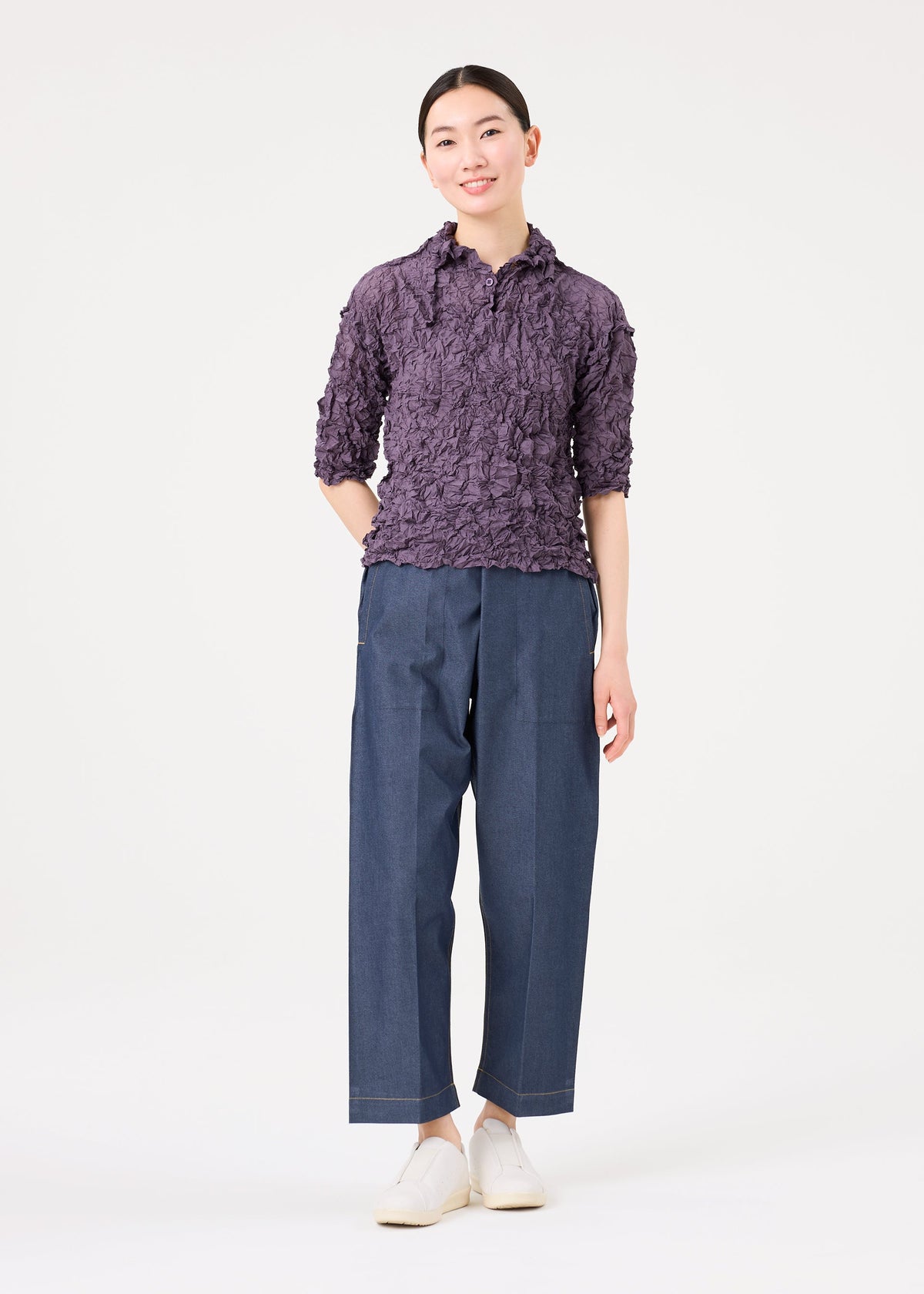 Zoomed view A person stands against a white background wearing the MERINGUE TOP by me ISSEY MIYAKE in purple, paired with wide-leg blue pants and white shoes, hands behind their back and a slight smile.