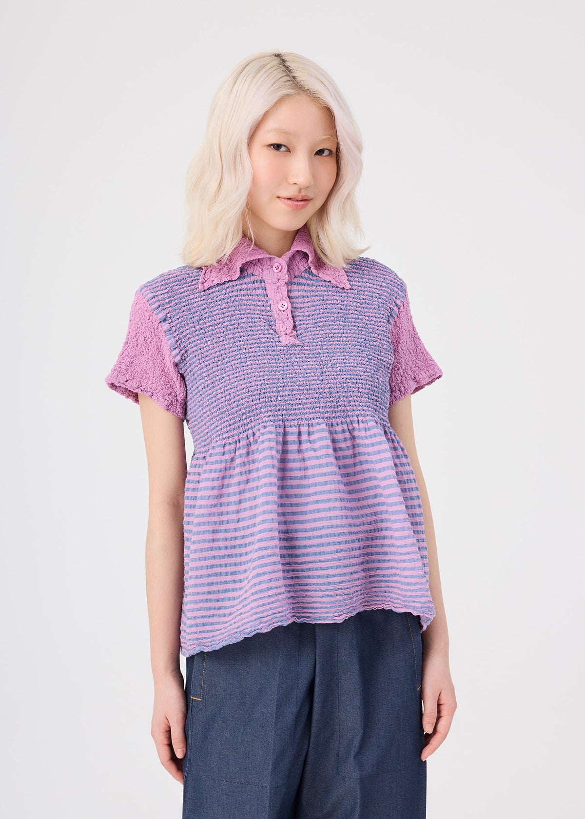 A woman with light blonde hair stands against a plain white background wearing the me ISSEY MIYAKE STRIPE CAULIFLOWER cotton short-sleeved top, featuring purple and lavender stripes with buttons, paired with blue pants. view 5 of 7