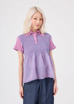 A woman with light blonde hair stands against a plain white background wearing the me ISSEY MIYAKE STRIPE CAULIFLOWER cotton short-sleeved top, featuring purple and lavender stripes with buttons, paired with blue pants.