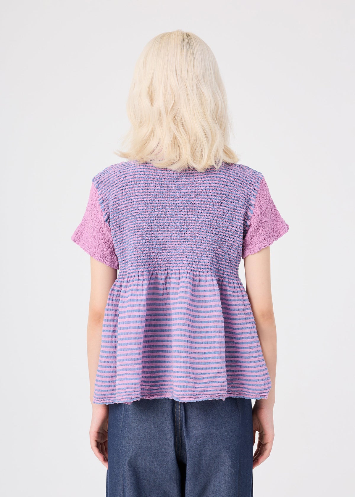 A person with shoulder-length blonde hair, seen from behind, wears the STRIPE CAULIFLOWER cotton short-sleeved top by me ISSEY MIYAKE—featuring purple and pink stripes and textured fabric—paired with blue pants against a plain light background. view 6 of 7