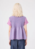 A person with shoulder-length blonde hair, seen from behind, wears the STRIPE CAULIFLOWER cotton short-sleeved top by me ISSEY MIYAKE—featuring purple and pink stripes and textured fabric—paired with blue pants against a plain light background.